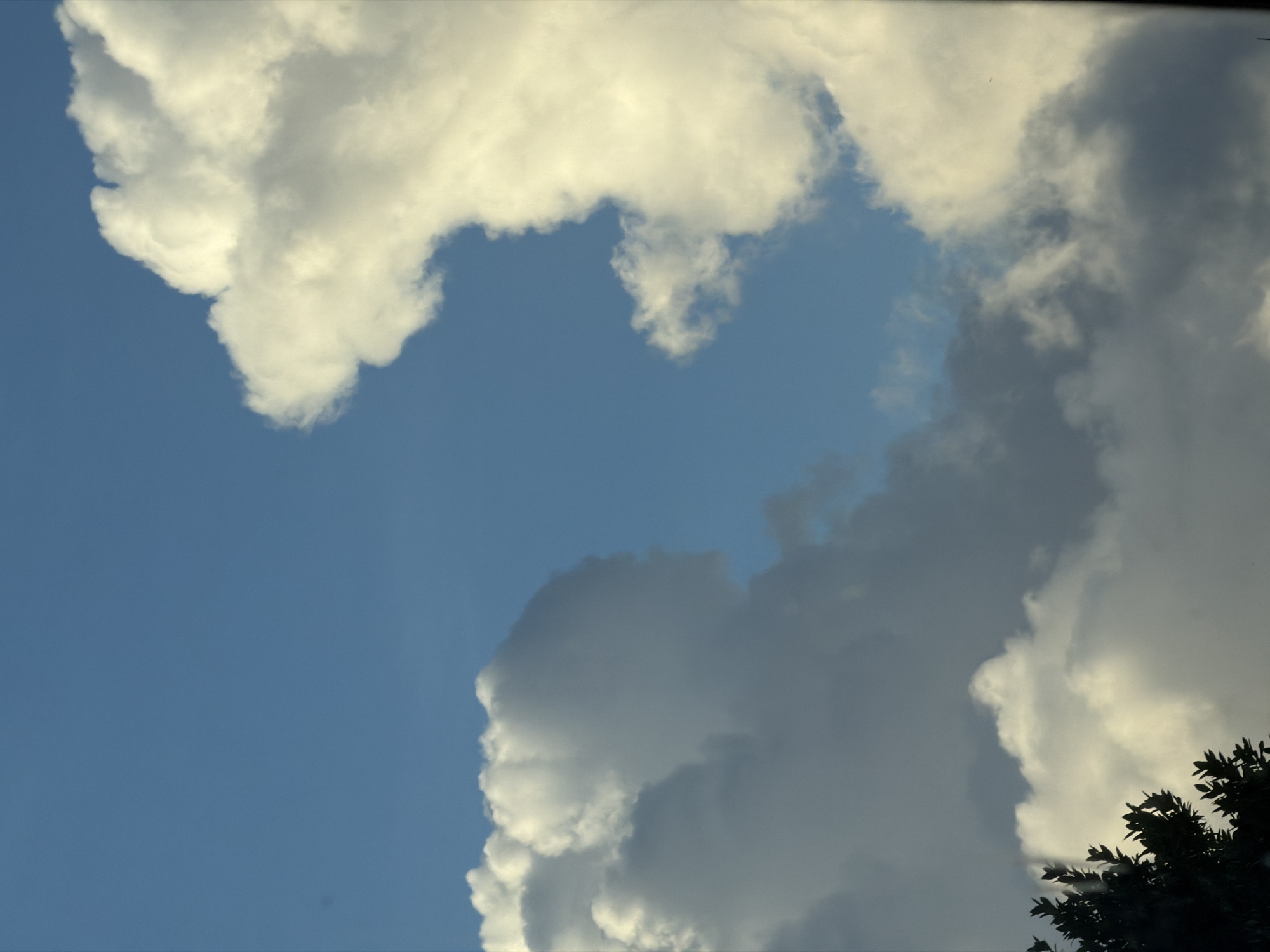 The image shows a section of the sky with cumulus clouds, partly covering the blue expanse. The lower right corner contains dark foliage. There is no indication this photo was taken in North Kuta, Indonesia.