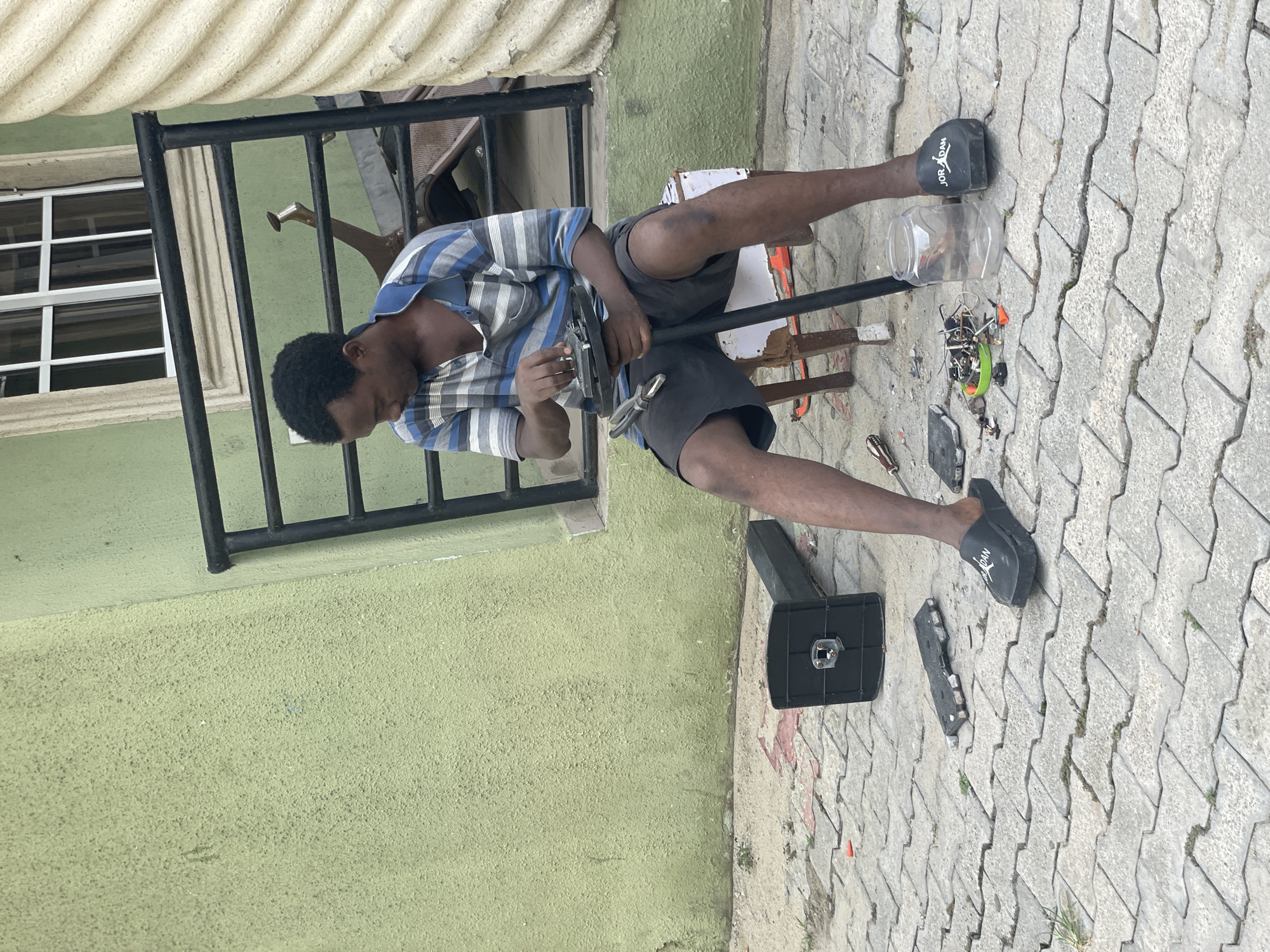 A person is seated on a wooden stool outdoors, working on what appears to be a small electrical device or appliance. The person is wearing a short-sleeved, striped shirt and dark shorts, along with black slides. Scattered around the person on the ground are various tools, components, and parts of the appliance. A clear plastic container sits near the person's feet. The environment is paved with interlocking stones. In the background, a green wall with a black metal railing and a window with white trim is visible. This scene is located in Ado, Nigeria.