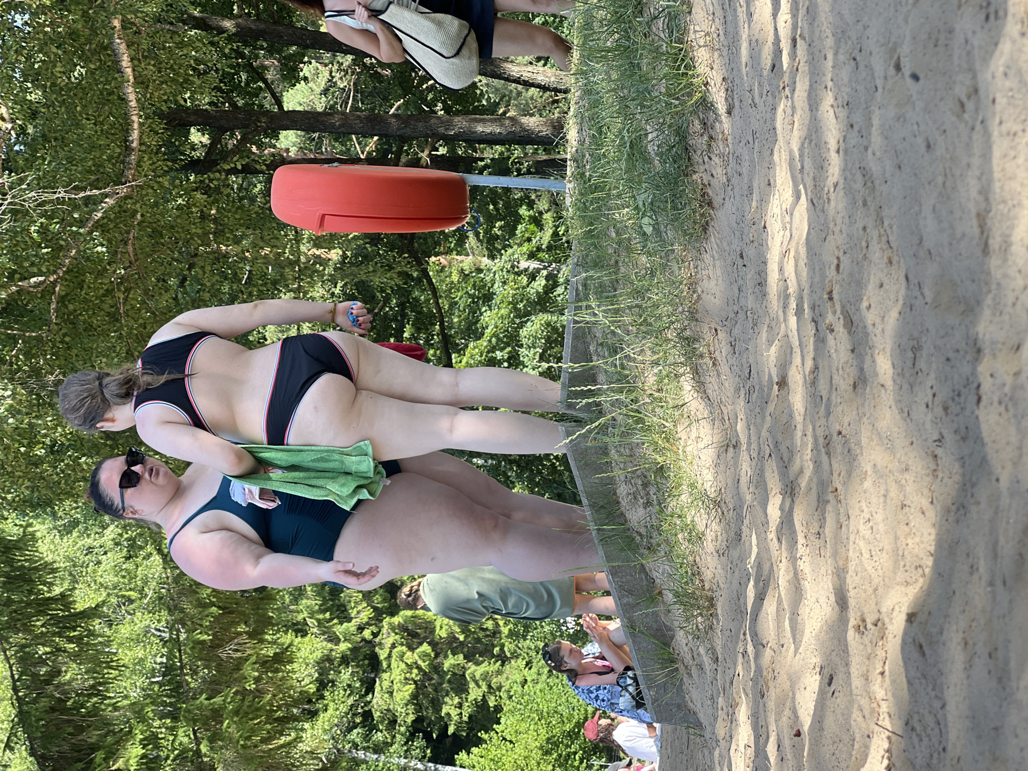 The image shows a group of people at a beach or park in Vilnius, Lithuania. The primary subjects are two women in swimsuits. One woman in the foreground is wearing a dark green one-piece swimsuit and sunglasses. She is lying on the sand, leaning on her arm. Another woman behind her is wearing a black and red striped bikini. She is also lying on the sand. A bright orange object, possibly a lifebuoy or a buoy, is visible in the background. The setting appears to be a sandy area with grass and trees, suggesting an outdoor recreational space. The lighting indicates it is daytime, likely sunny. The overall mood seems relaxed, consistent with a day spent outdoors at a beach or park. There is no visible text in the image.