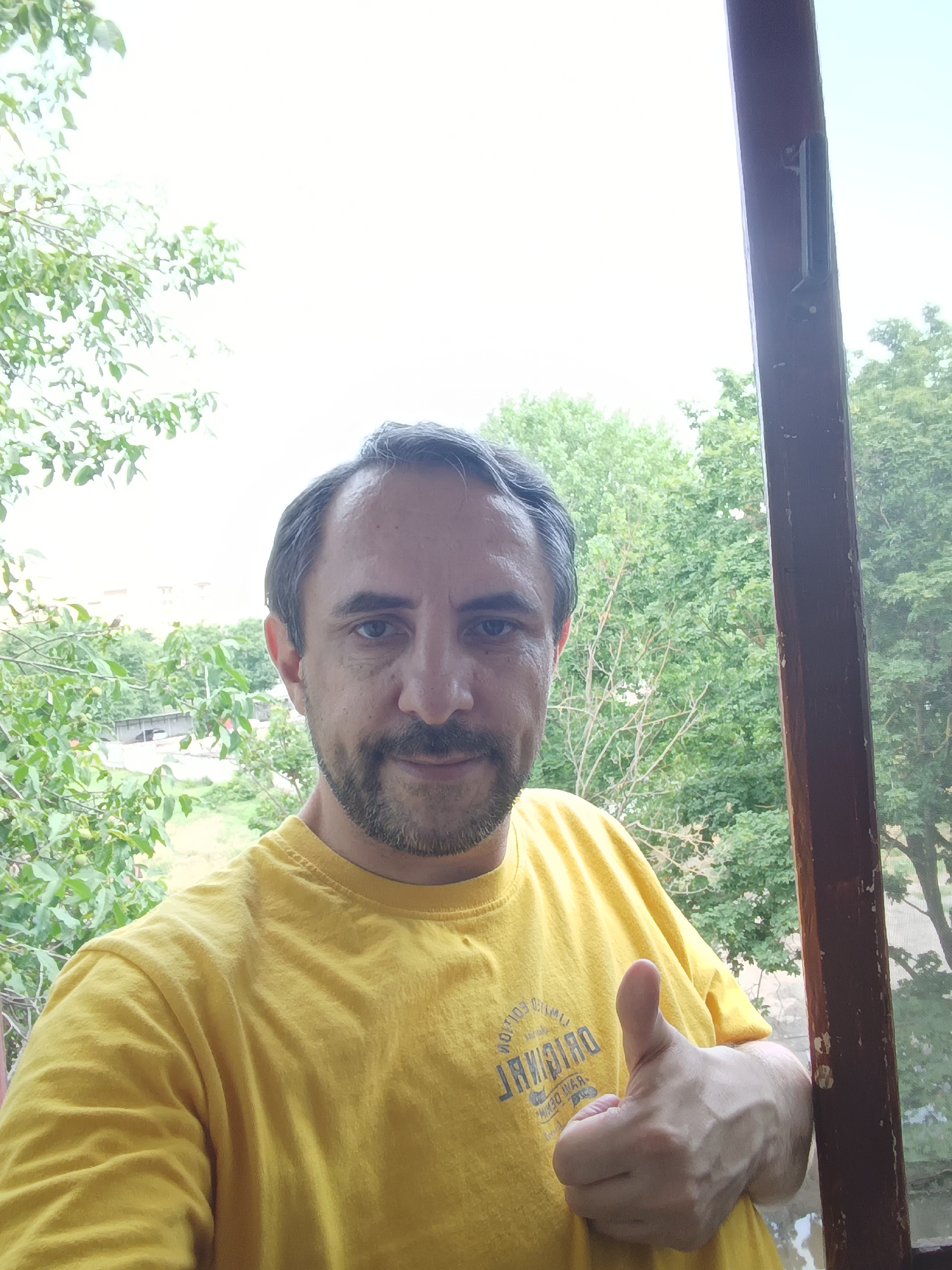 In Izmail, Ukraine, a man with short, dark hair, a mustache, and a beard is pictured giving a thumbs-up. He is wearing a bright yellow t-shirt with a faded print. The man is standing on a balcony or porch, as indicated by the wooden frame visible on the right side of the image. Beyond him, lush green trees and foliage are abundant, suggesting a setting surrounded by nature. The sky is overcast.