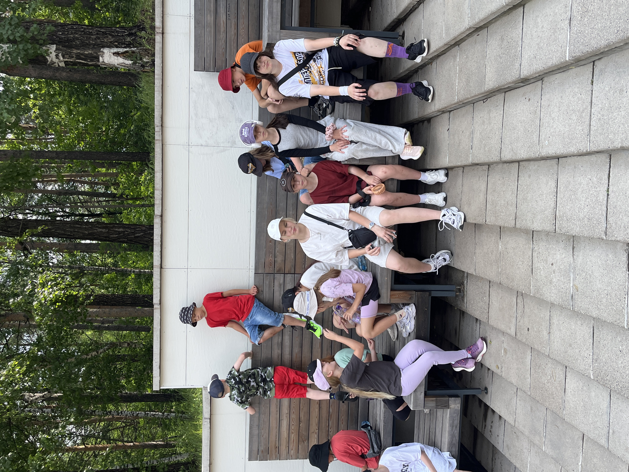A group of ten young people, appearing to be children and teenagers, are gathered on wooden benches and concrete steps in an outdoor urban park setting. The location is identified as Divnogorsk, Russia, with a backdrop of lush green trees and some birch trees. The group is diverse in their attire, with various t-shirts, shorts, pants, and hats.
On the upper bench, a boy in a red t-shirt and denim shorts is seated on the far right. To his left, a person in an orange t-shirt and red bucket hat is also seated. Next to them, a group of four individuals are seated. From left to right, a person in a black t-shirt and blue jeans, a person wearing a white bucket hat and a grey and black top, and a person in a grey tracksuit are visible. The furthest left on this bench is a person in a red t-shirt and black shorts.
On the lower bench and steps, there are four more individuals. On the left, a boy in a camouflage t-shirt and red shorts is climbing on the bench. To his right, a girl in a dark t-shirt and light purple pants is seated, reaching out towards another girl. The second girl is wearing a light purple t-shirt and dark shorts, holding a water bottle. On the far right of the lower seating area, a person in a white t-shirt and light grey shorts is seated. Further to the right and on the steps, a person is visible wearing a white t-shirt and black shorts, with distinct purple socks.
The environment appears to be a modern park with clean lines, featuring wooden slatted benches and concrete tiered steps. The lighting suggests it is daytime with sunlight filtering through the trees. The overall atmosphere seems relaxed and casual, with the group interacting and posing for a photograph.