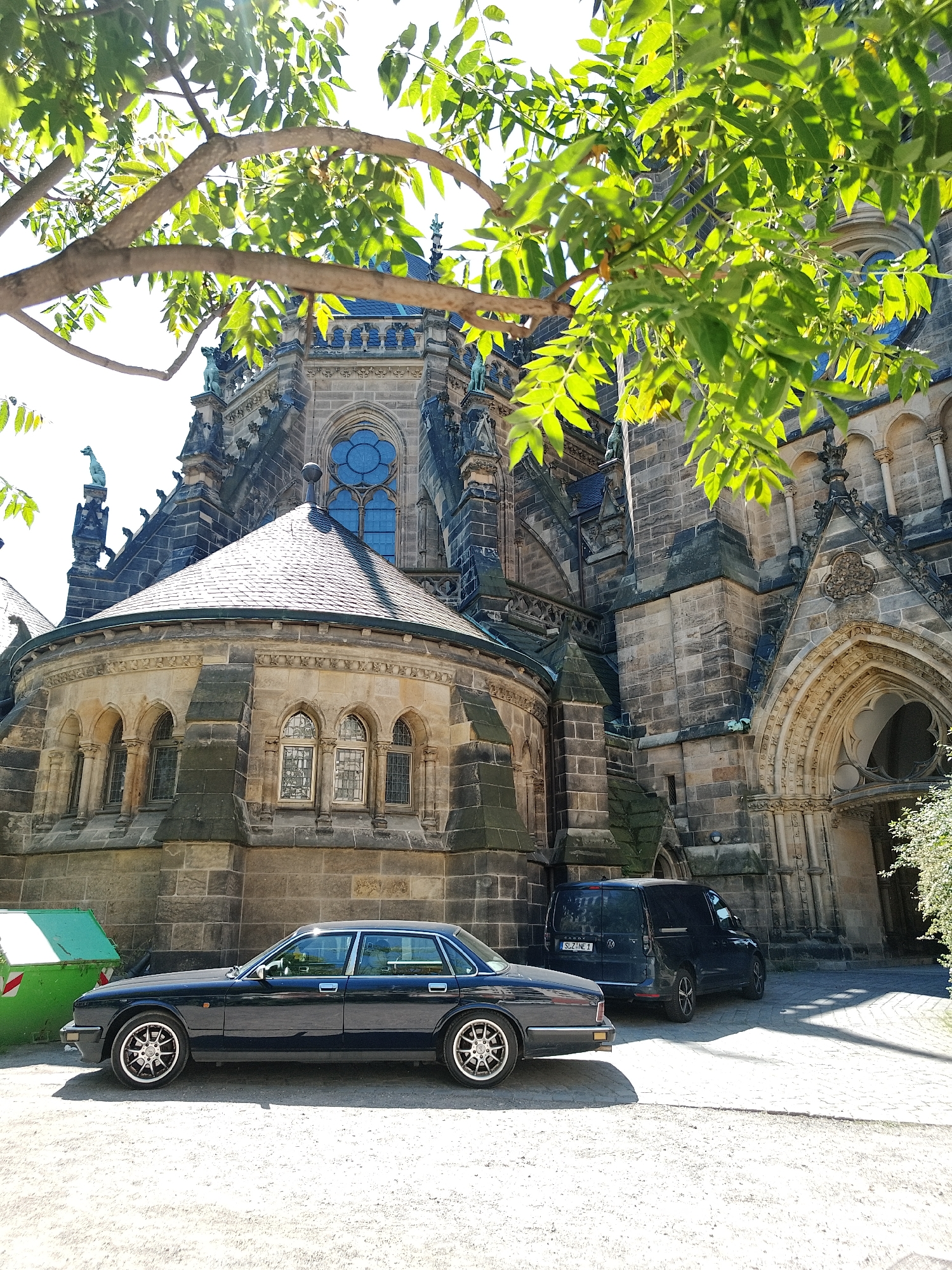 The image depicts a large stone building, potentially a cathedral, located in Unknown, Unknown. The architecture is ornate, with gothic elements such as pointed arches, detailed carvings, and a circular window. A section of the building has a rounded apse-like structure with several windows. The building appears to be constructed of gray stone blocks. In the foreground, there is a black sedan-style car parked along the side of the building. Behind it, there is a dark-colored van. A green dumpster is visible on the left side of the car. A tree branch with green leaves partially obstructs the view of the building in the upper portion of the image. The ground is a mix of dirt and gravel, and there is a paved area near the entrance to the building. The scene appears to be outdoors and in daylight.