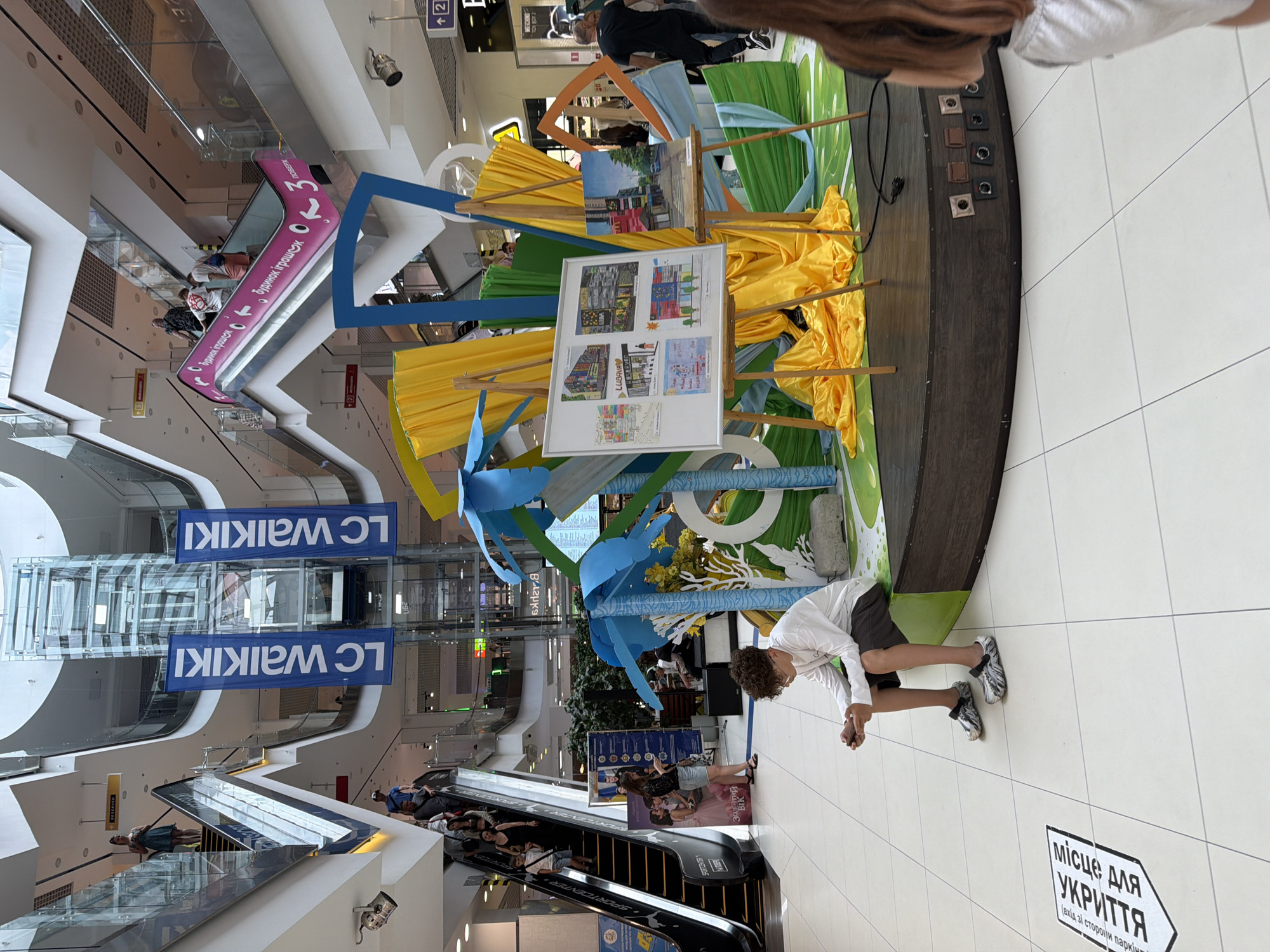 The image captures an interior scene within a multi-level shopping mall, likely in Cherkasy, Ukraine, given the Cyrillic text visible on signage and floor markings. In the foreground, a young male with curly hair, wearing glasses, a white long-sleeved shirt, and dark shorts, sits on a curved, dark wooden platform, looking down at a mobile device in his hands. He is wearing patterned clogs. The platform itself features a display with easels holding artwork. One easel supports a whiteboard showcasing multiple small drawings and text, while another displays a framed painting of a street scene. Behind the artwork, vibrant yellow and green fabric drapes create a backdrop, interspersed with blue and white cylindrical structures. The floor is tiled with a white pattern. To the left of the platform, a floor marking with Cyrillic text indicates a shelter. In the mid-ground, escalators are visible, with people ascending and descending. One escalator has a pink handrail with Cyrillic text and numbers. Banners with the "LC Waikiki" logo hang vertically near a glass elevator shaft. The upper levels of the mall are visible, with shops and more escalators. In the far right background, a man in a black t-shirt and dark pants walks away from the camera. The overall environment is brightly lit, indicative of an indoor commercial space.