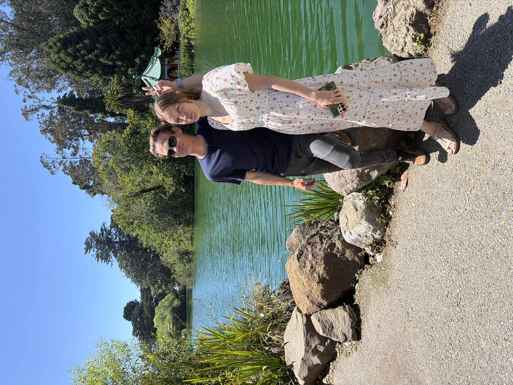 A young Caucasian couple stands on a paved path next to a calm, green lake with a Japanese-style pagoda visible in the background. The man, on the left, wears sunglasses, a navy blue t-shirt, and dark, fitted jeans with a motocross-inspired design on the thighs. He has light brown hair and is looking to his left. The woman, on the right, has blonde hair pulled back and is wearing a white, floral print maxi dress with puff sleeves. She is holding a phone in her right hand and making a peace sign with her left hand. She is looking at the camera. The lake is surrounded by lush green trees and vegetation. The path is made of gravel and there are large rocks on the left side of the image. The scene appears to be in a park setting on a sunny day with a clear blue sky. This scene is likely the Japanese Tea Garden in Golden Gate Park, San Francisco, California, United States of America, given the architecture of the pagoda and the surrounding landscape.