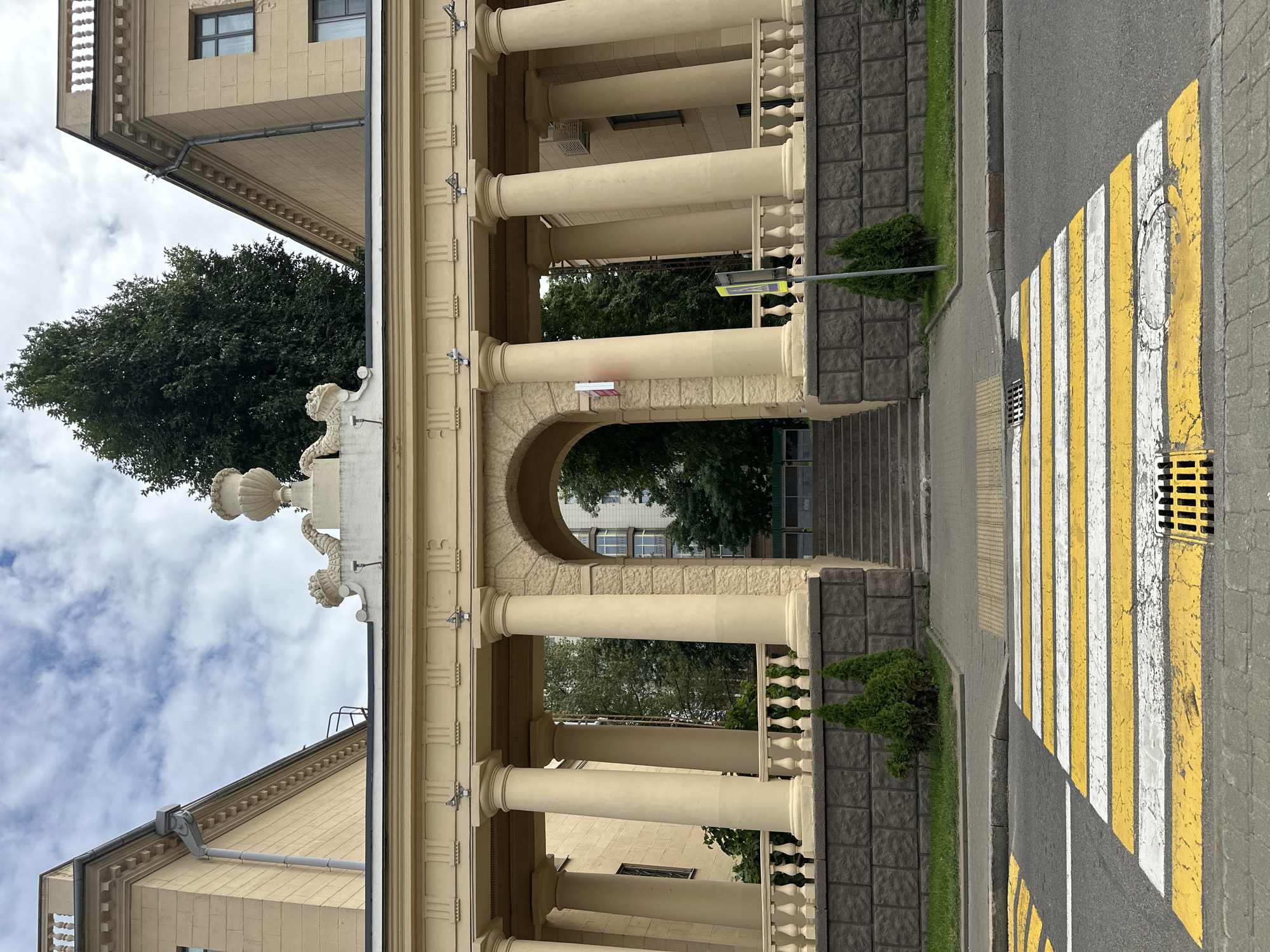 The image shows a pedestrian crossing with yellow and white stripes in front of a light yellow building with classical architectural elements. The building has a colonnade with Corinthian columns and a decorative urn on top. There are two green cypress trees planted in front of the wall. The sky is cloudy. The scene appears to be a city street in Minsk, Belarus.