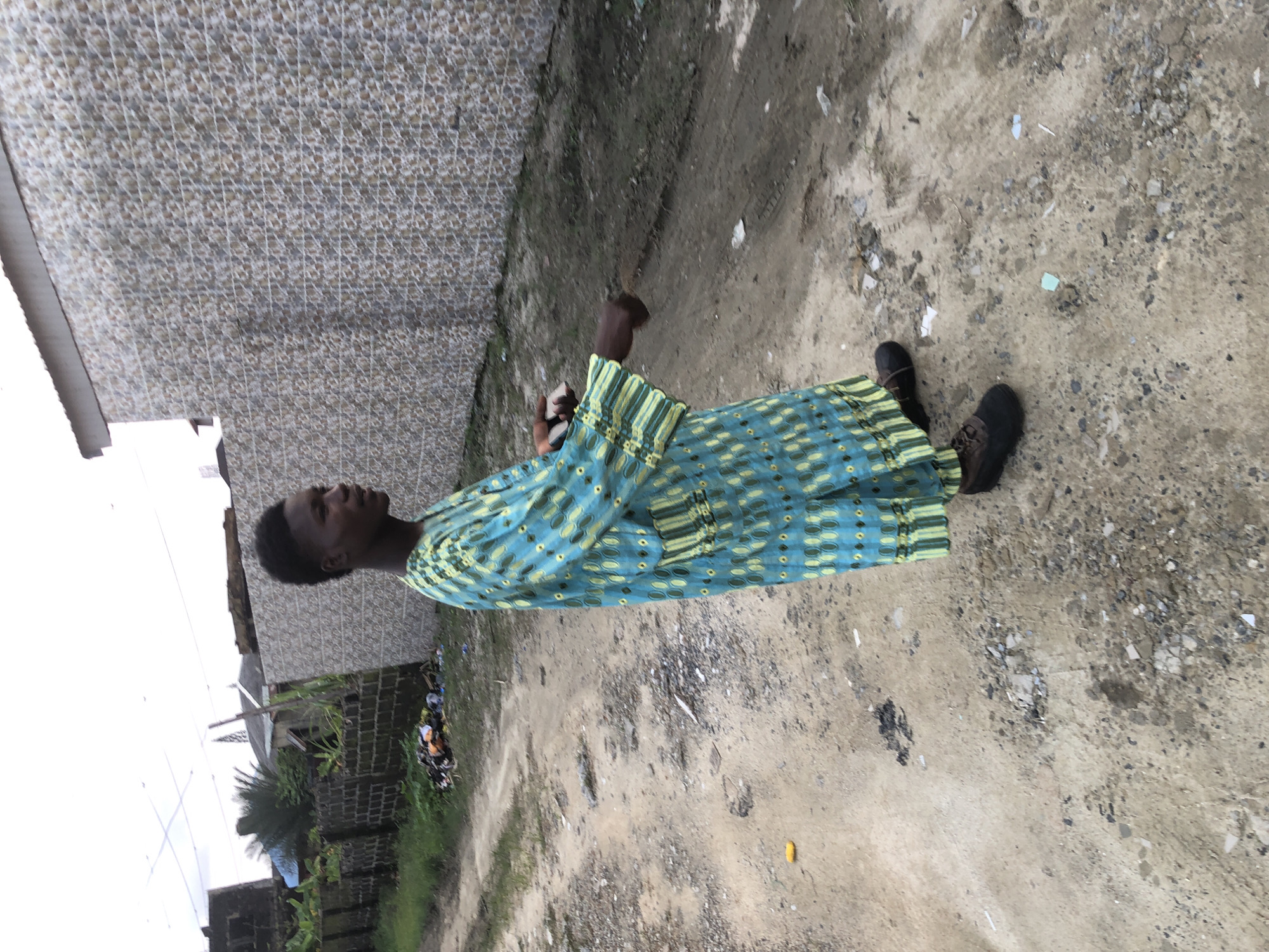 A young Black male stands on a dirt path in front of a patterned tiled wall. He is wearing a long, blue and green patterned robe and dark shoes. The robe has a repeating circular pattern in a lighter shade of blue and yellow accents at the cuffs and hem. The path is made of dirt and gravel, with some scattered debris and patches of dry grass. In the background, there are brick walls, vegetation, and buildings. The lighting suggests an overcast day. The location is identified as Ado, Nigeria.