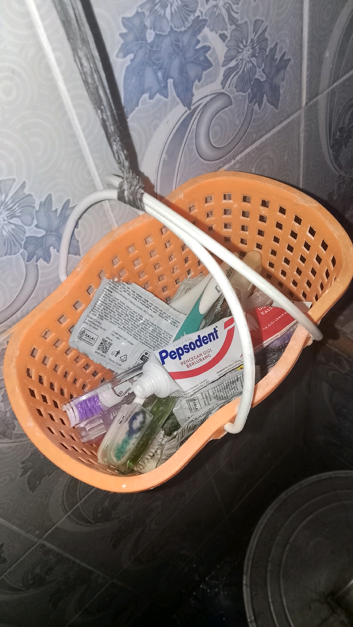 An orange plastic basket hangs by a rope. The basket contains dental hygiene products. A tube of Pepsodent toothpaste is visible, along with a toothbrush and what appears to be a travel-sized mouthwash bottle. The background shows a tiled wall with a floral pattern. The floor appears to be dark tiled. The scene is likely in a bathroom. The location is specified as Mulyo Haji, Indonesia.