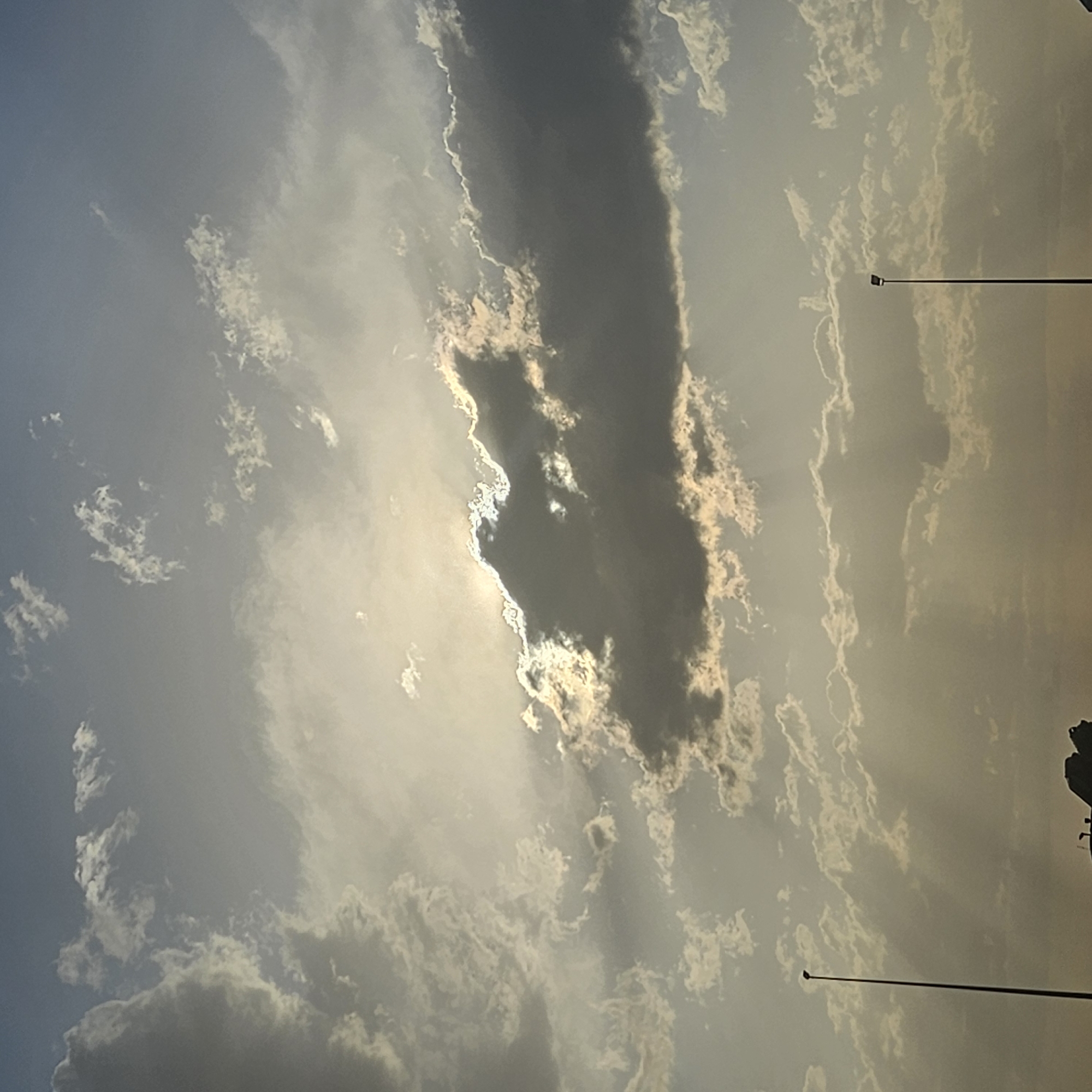 This is a photograph of a cloudy sky over Algora, Spain. The sun is partially obscured by a large, dark cloud in the center of the frame, with rays of light filtering through the edges. The sky is a mix of light blue and hazy grey, with scattered white clouds of various sizes and shapes. In the foreground, silhouetted against the sky, are two tall streetlights on either side of the image. The lower portion of the image shows a faint silhouette of structures and possibly some vegetation. The overall lighting suggests it is either sunrise or sunset.