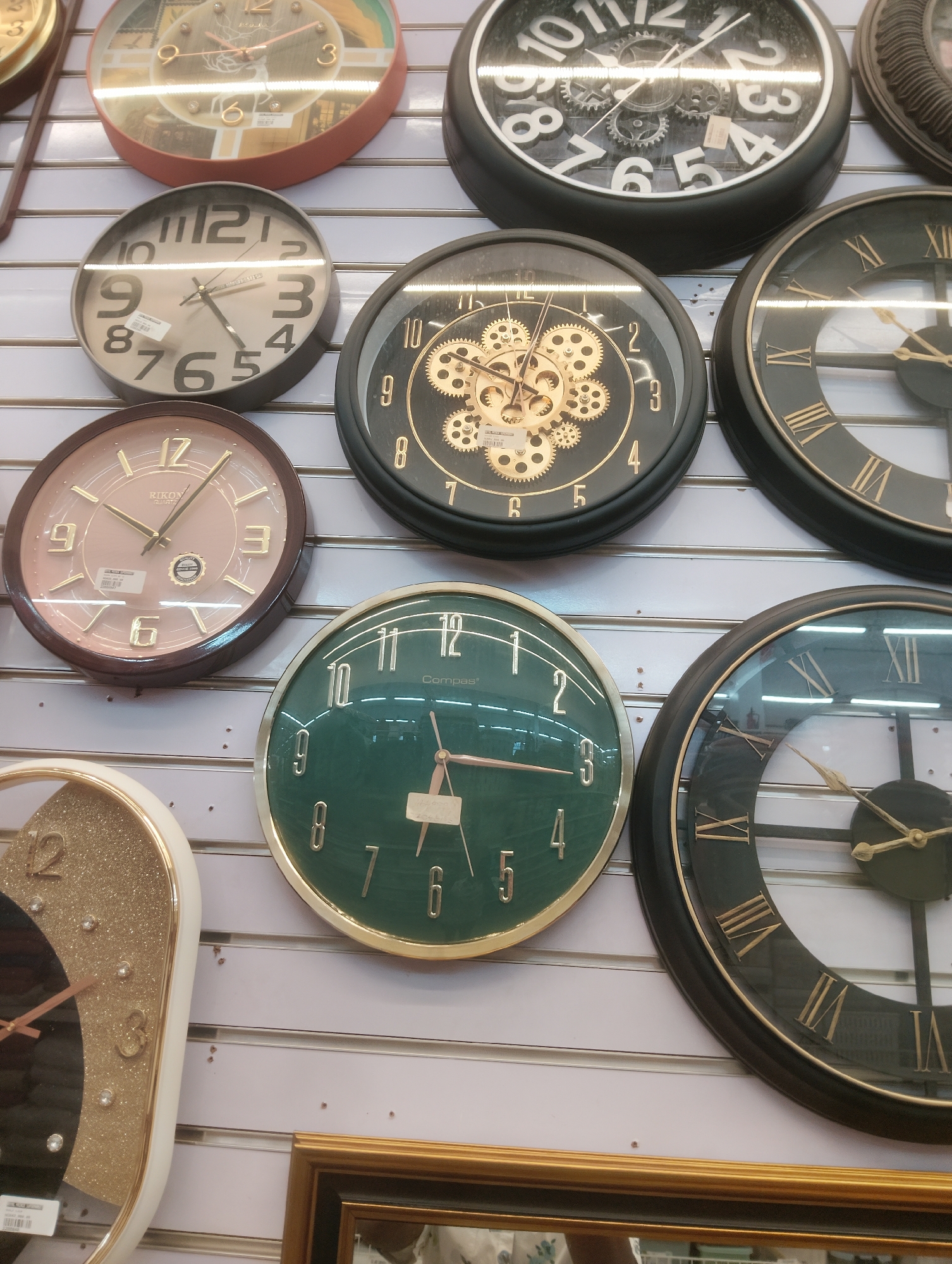 The image shows a wall adorned with an assortment of clocks, primarily displayed in a retail setting, potentially a market stall or a shop. The clocks are of various designs, sizes, and colors. Some feature Arabic numerals, while others display Roman numerals or decorative gears. Several clocks are round, while one appears to be an oval shape. The wall itself is paneled with horizontal strips. The lower portion of the image includes a glimpse of a framed picture or mirror, suggesting the presence of other items for sale or decoration. The overall scene is that of a commercial establishment showcasing timepieces. The location of Aketu-Oja, Nigeria is not discernible from the image itself, but the context suggests a commercial area within that city.