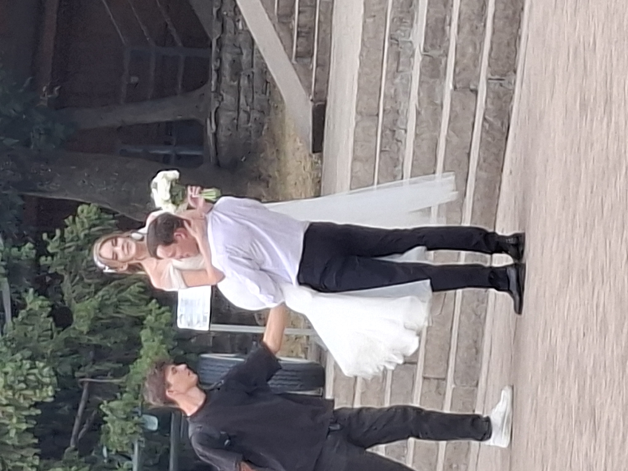A wedding scene unfolds outdoors, featuring a bride and groom in formal attire, and another man dressed casually. The bride, adorned in a white off-the-shoulder wedding dress and a delicate headband, is being carried by the groom, who is in a white dress shirt and black trousers. He holds a bouquet of white flowers in his left hand. The other man, wearing a black t-shirt and dark trousers, stands to their left, appearing to interact with the groom. They are positioned on a paved surface with stone steps leading up behind them, suggesting a public outdoor location. Trees with lush green foliage form the backdrop. The lighting indicates it is daytime, possibly with overcast conditions. The scene is captured from a medium distance, with the focus on the people. There is no identifiable information to confirm the location as Almaty, Kazakhstan.