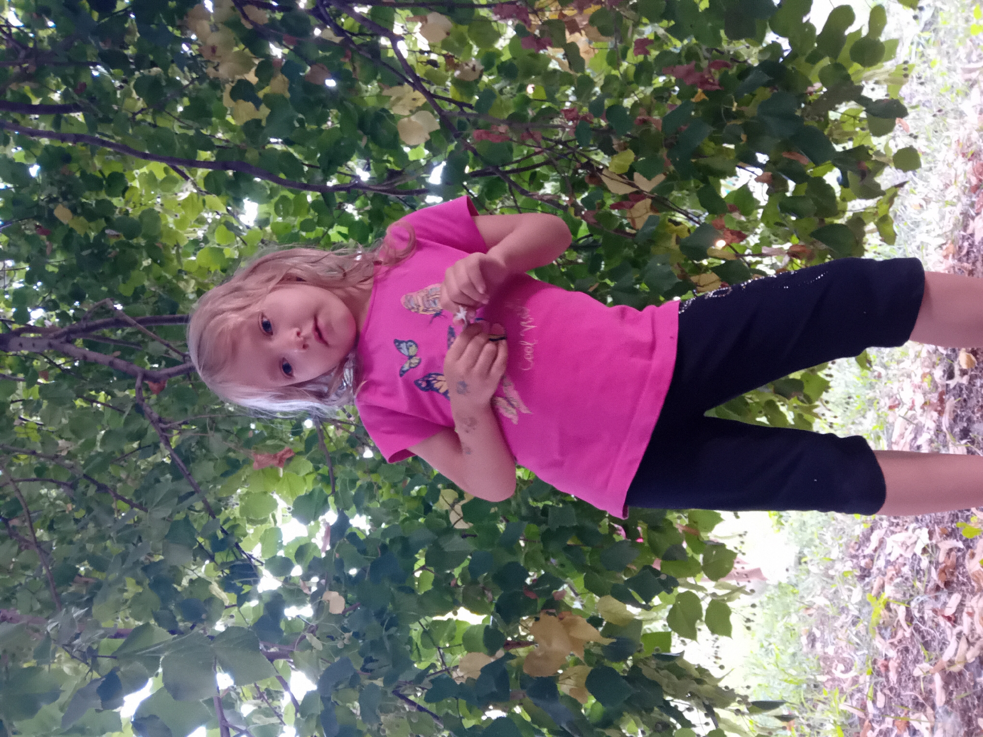 A young girl with blonde hair and a pink t-shirt stands in front of a tree with green leaves. She is wearing black pants. The girl appears to be outdoors, with foliage and some dry leaves on the ground. The image does not contain enough information to determine the specific location in Chuhuiv, Ukraine.