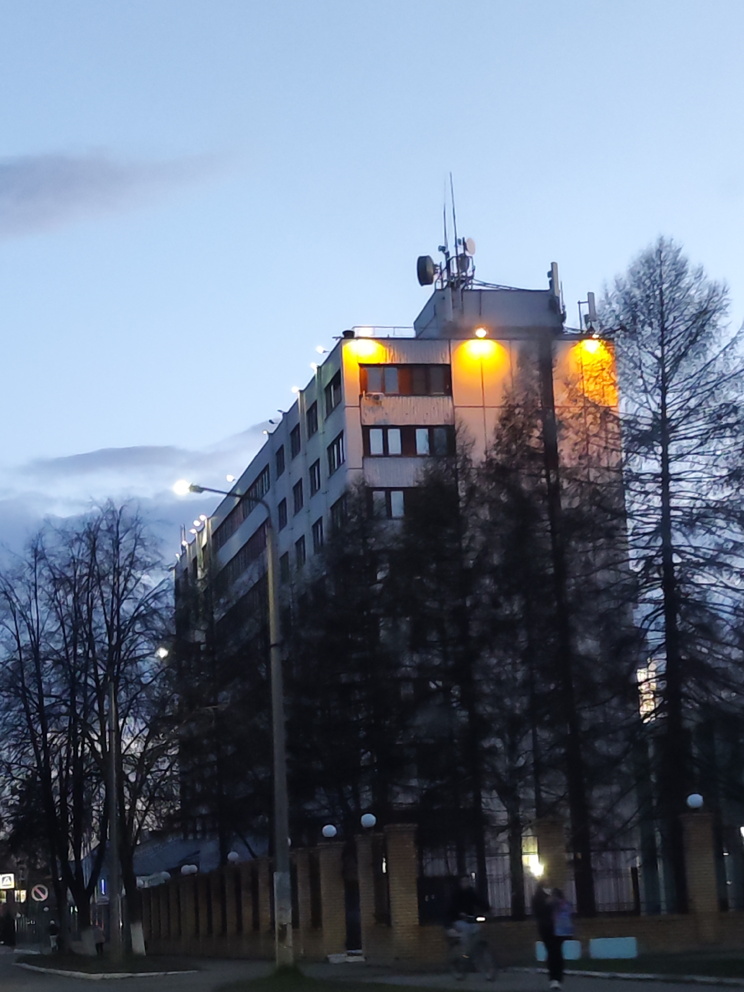 The image depicts an urban or semi-urban scene at dusk or early evening, characterized by the onset of artificial lighting against a pale blue sky with soft, diffuse clouds.
A prominent multi-story building, likely an apartment complex or administrative block with a functional design, dominates the center. Its facade is illuminated by a series of bright, warm yellow-orange lights positioned along the top edge, and multiple windows are visible across its stories. The flat roof of the building hosts several antennas, including what appear to be cellular communication antennas and possibly a satellite dish.
The building is flanked and partially obscured by trees. On the right, dense evergreen trees stand tall, their dark forms contrasting with the illuminated building. On the left and in the foreground, bare deciduous trees with intricate branch patterns line what appears to be a street or pathway. A tall streetlight positioned on the left side of the building emits a bright, white light.
In the middle ground, a brick fence or wall spans horizontally, featuring evenly spaced round, white light fixtures along its top. The foreground shows a paved surface, likely a road or sidewalk. Two blurry figures are visible in the lower right corner near the fence: one person appears to be riding a bicycle, and another is walking alongside.
On the far left, a road sign is visible, displaying a circular "no entry" or "no vehicles" symbol, but any textual information on it or other nearby signs is not decipherable due to blurriness or distance. The scene conveys a calm, transitional period from day to night, common in a residential or mixed-use area in a location like Chaikovsky, Russia.