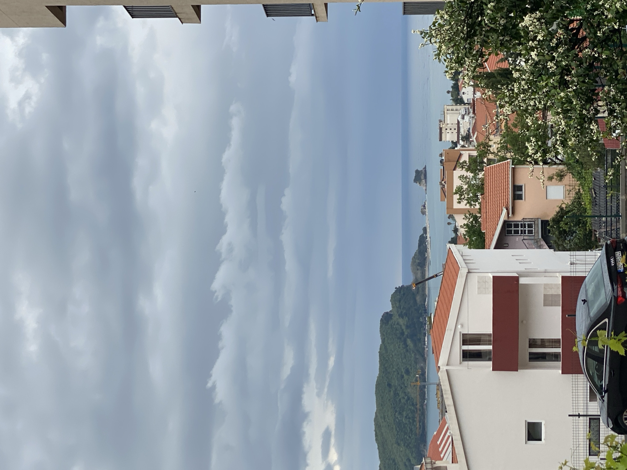 This landscape-oriented image captures a scenic view of a coastal town, consistent with the location context of Budva, Montenegro, from an elevated perspective.
In the immediate foreground, the top of a modern building's overhang, featuring horizontal slats, frames the upper edge of the image. Below this, a cluster of residential buildings with predominantly white facades and warm reddish-brown terracotta roofs are visible, interspersed with lush green foliage and trees bearing small white blossoms. One prominent building in the mid-right features distinct dark red/maroon panels. A black car, identifiable as an Audi by its emblem on the license plate holder, is partially visible on the right, with its rear license plate clearly showing "BD 89241".
Beyond the urban settlement, a large, verdant mountain or hill, thickly covered in trees, slopes down to meet a calm, blue-grey sea. A yellow construction crane with partially visible text "CONSTRU..." (likely "CONSTRUCTION") is situated on the hillside near the water. Several small, rocky islets dot the tranquil water. The sea extends into the distance, merging with a horizon under a heavily overcast sky. The sky is dominated by layers of soft grey clouds, suggesting a cloudy daytime, possibly without direct sunlight. No people or overt activities are visible, but the urban setting and crane imply ongoing life and development.