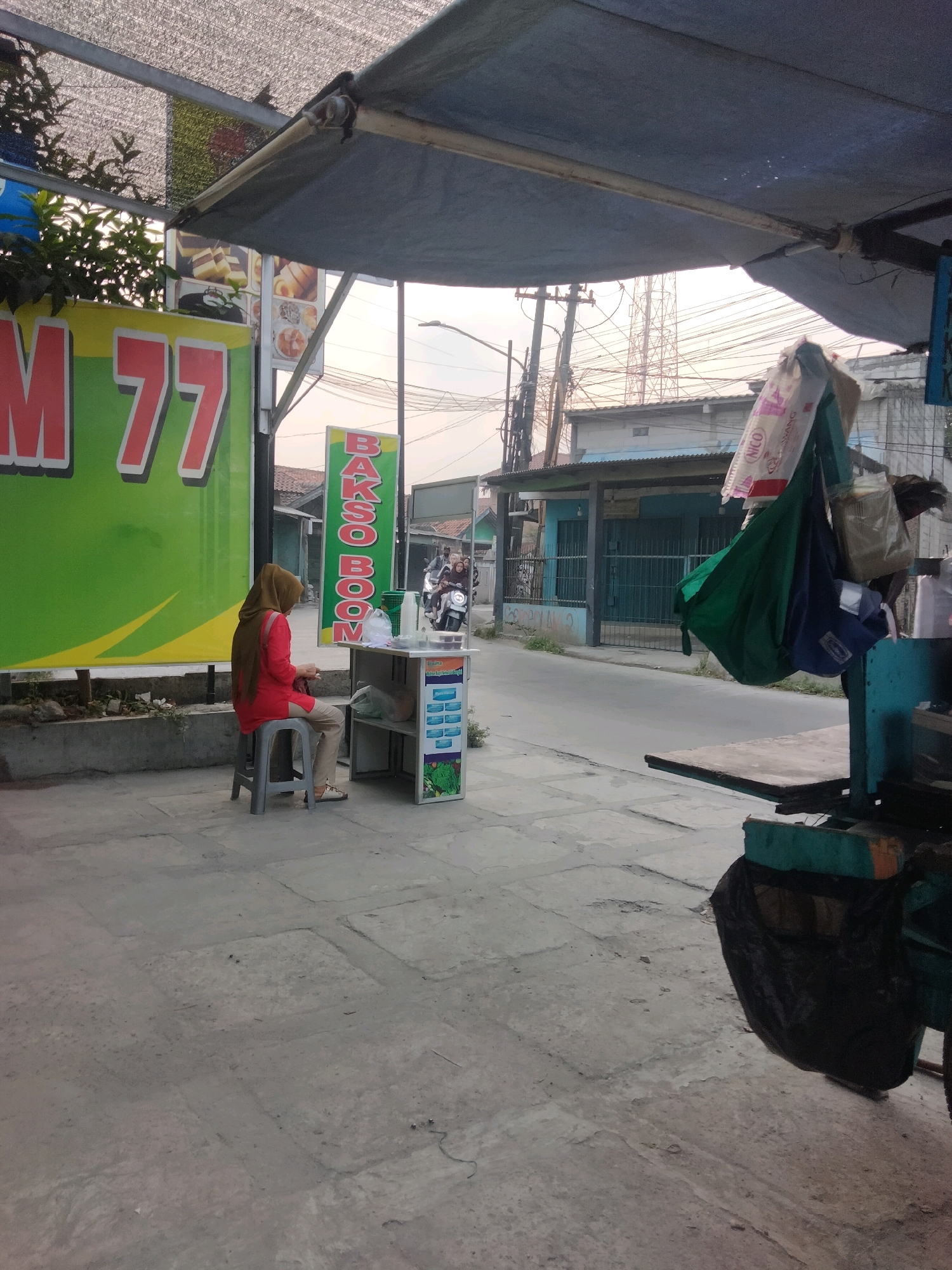 The image depicts a street scene in Pisangan Jaya, Indonesia. On the left, a vibrant green banner with red lettering that reads "M 77" is visible. In front of the banner, a woman wearing a hijab and a red top is seated on a grey stool. She is facing away from the camera, looking towards a small food stall. The stall has a green sign with white lettering that spells "BAKSO BOOM." The stall is equipped with shelves containing various items, including a plastic bag and what appear to be food containers. Behind the stall, a street is visible with buildings and a few motorcycles. In the background, utility poles and electrical wires are prominent, and a distant telecommunications tower can be seen. The ground is paved with concrete tiles. Several bags, including a green and a dark blue one, are hanging from the stall's structure. A black trash bag is attached to the lower part of the stall. The sky is overcast, suggesting it might be early morning or late afternoon.