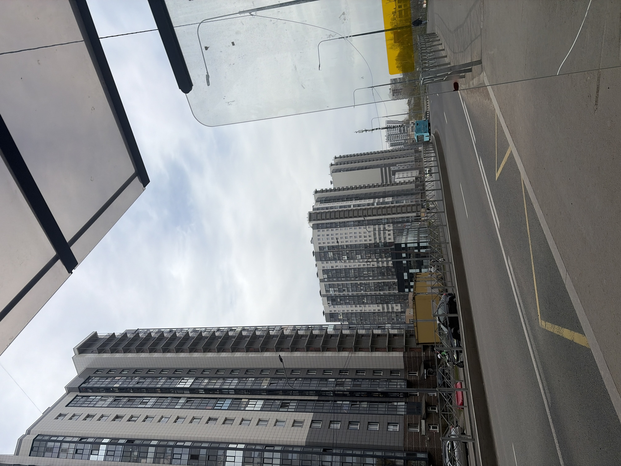 The image depicts a street scene in what appears to be Saint Petersburg, Russia. High-rise apartment buildings dominate the background, their facades a mix of concrete, glass, and dark accents. A multi-lane road runs alongside the buildings, with yellow markings indicating bus stops or restricted parking zones. A bus shelter structure is visible in the foreground, partially obscuring the sky. Several parked cars line the street next to the bus stop. The sky is overcast.