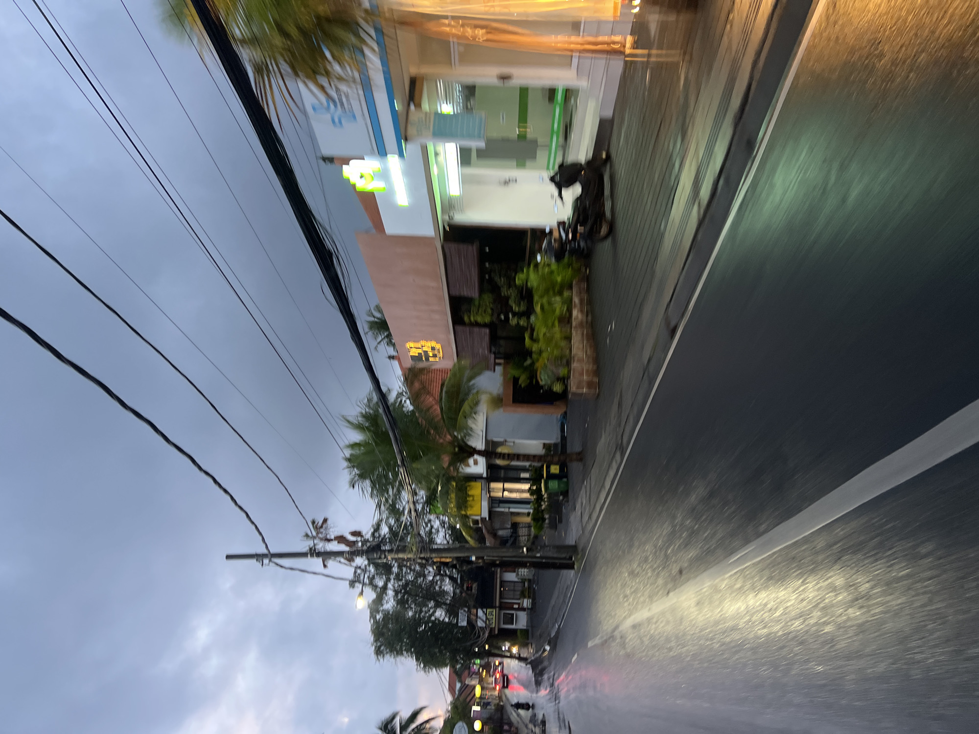 The image shows a street in North Kuta, Indonesia, at dusk or dawn. The sky is overcast and grey, with dark clouds. The street is wet, reflecting the lights from the buildings and vehicles.
On the right side of the street, there are several buildings with storefronts. One building has a sign with a blue cross and what appears to be a logo. Another building has a sign with yellow lettering. A parked motorcycle is visible in front of one of the buildings.
On the left side of the street, there are more buildings, some with illuminated signs. A few cars and a motorcycle are seen driving on the street, their lights blurred due to the motion. Trees and palm trees are scattered along the street.
Utility poles and numerous electrical wires are prominent in the sky above the street. The overall scene depicts a wet, dimly lit urban environment in Indonesia.