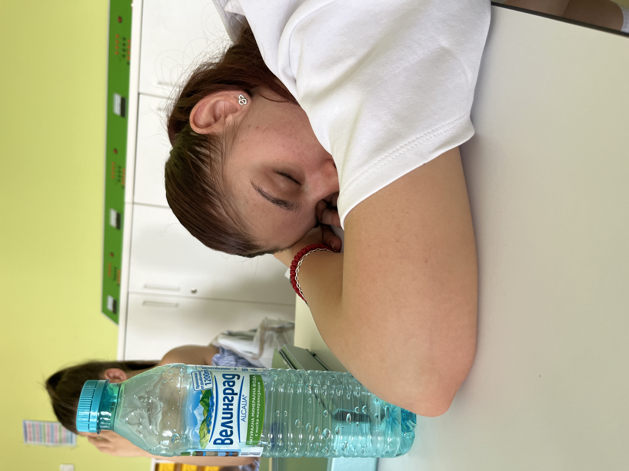A young woman is resting her head on a white surface, possibly a desk. She is wearing a white shirt and has brown hair pulled back. A water bottle is in the foreground, labeled "Велинград" (Velingrad) in Cyrillic script. The bottle's label also mentions it is natural mineral water with low mineralization and that it originates from a depth of 1200m. The location is identified on the bottle as Velingrad, Bulgaria, NOT Plovdiv. The background includes a green wall and white cabinets. A red bracelet adorns her wrist, and a small earring is visible in her ear.