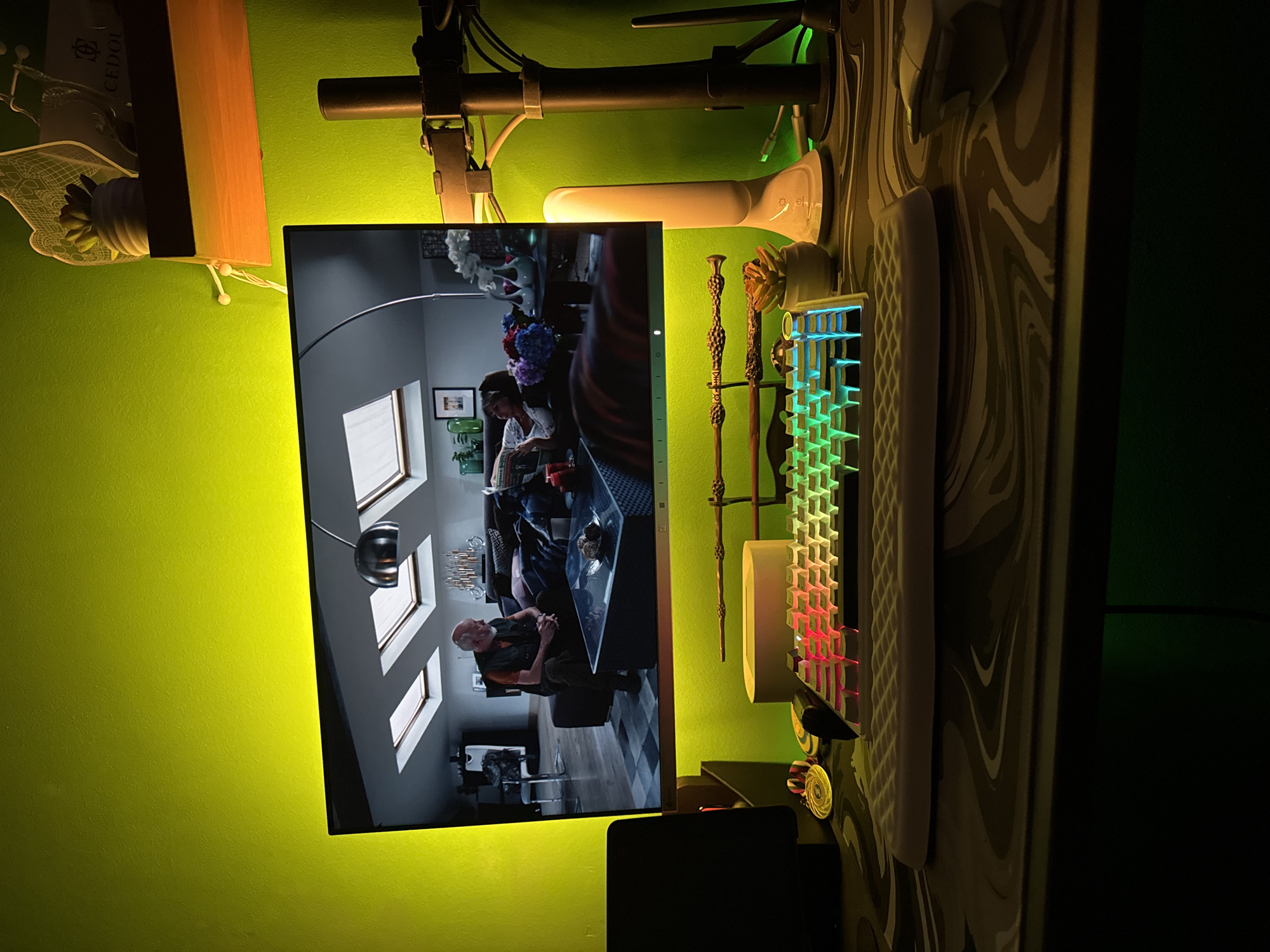 A monitor displays a scene from the film "Harry Potter," depicting an interior setting. The monitor is situated on a desk, adjacent to a keyboard, possibly a mechanical keyboard based on its design, and a decorative, possibly Harry Potter-themed, wand stand. The background reveals a vibrant green wall illuminated by ambient lighting. Cables and a scooter steering column are visible above the monitor. The desk surface has a patterned design. The monitor is the focal point, suggesting someone is watching "Harry Potter." The location, Varna, Bulgaria, cannot be determined from the image.