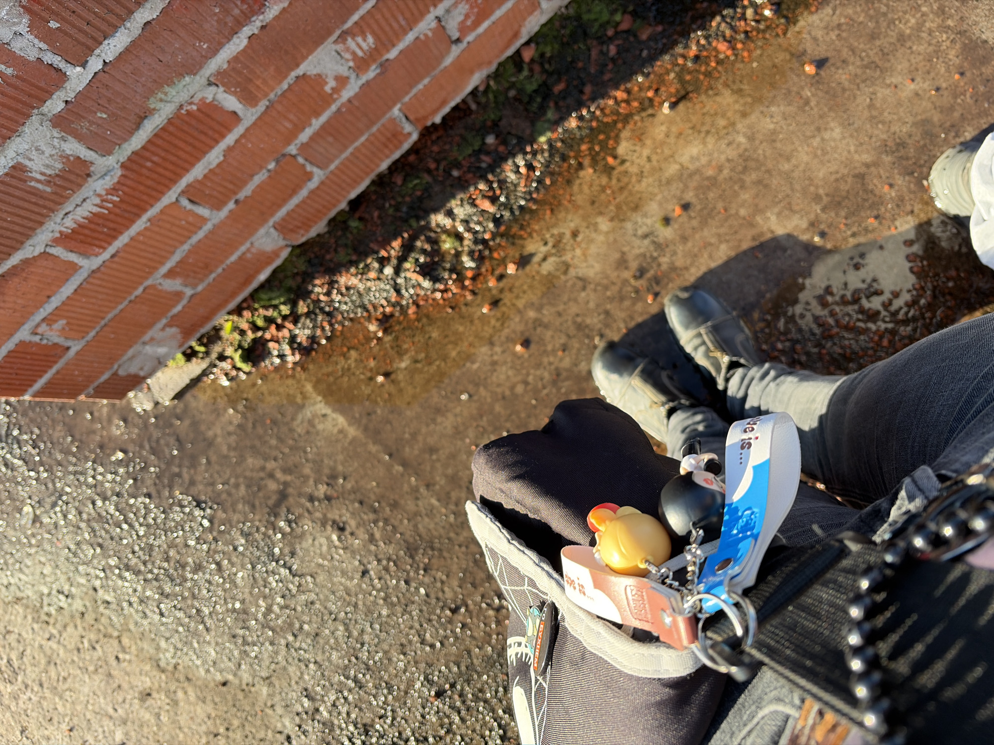 A high-angle outdoor shot shows a person sitting on a paved surface next to a brick wall. The ground appears wet and reflects the sunlight. The person is wearing dark pants and boots. Several keychains hang from a black bag or pocket, including a yellow duck, a small figurine, and a blue and white tag that reads "We is...". The brick wall is orange-red and shows signs of construction with visible mortar. The lower right of the image shows the top of the person's white shoe. The scene is devoid of other people and suggests an urban environment.