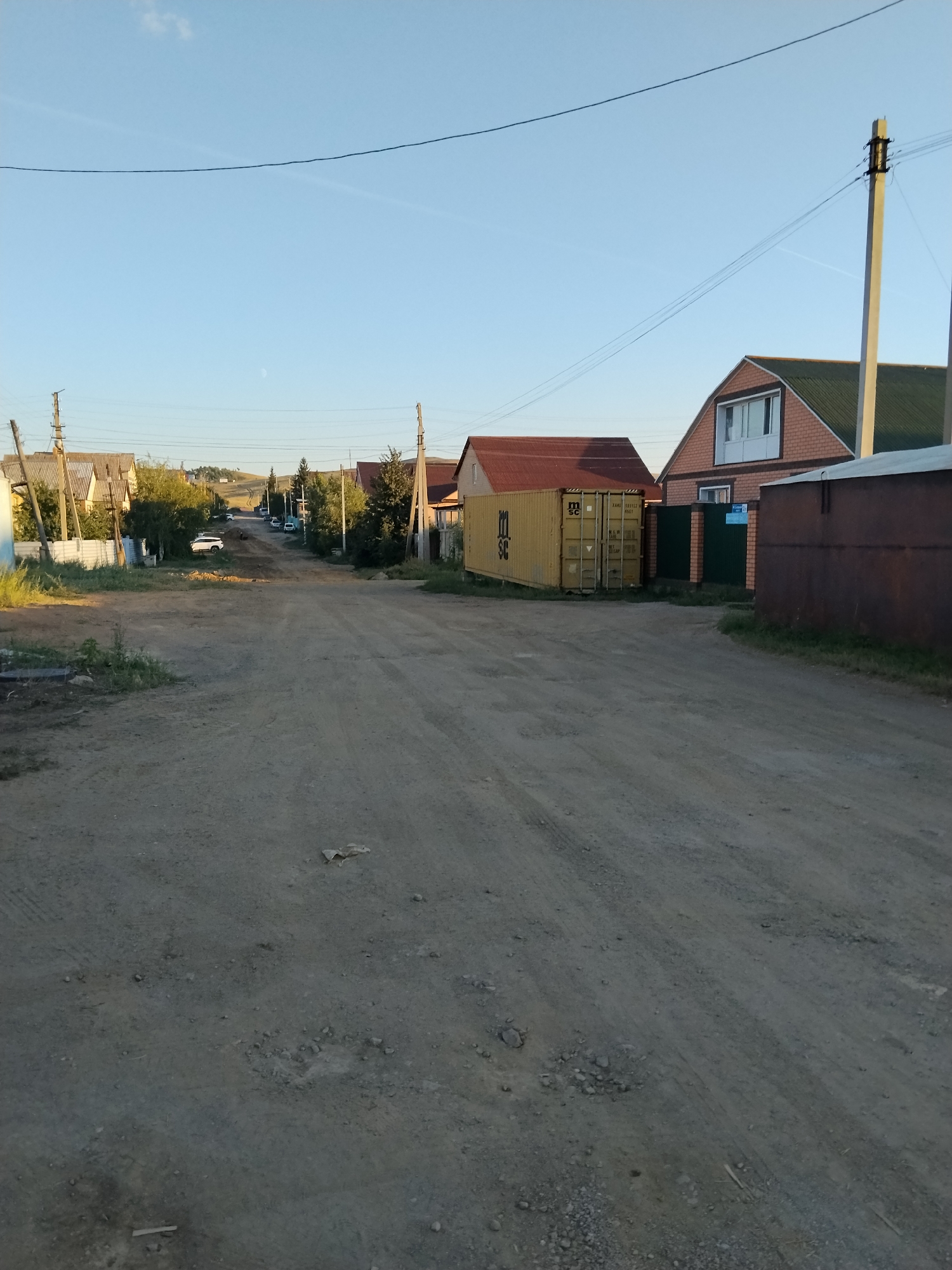 The image depicts a dirt road in a rural or suburban area. On the left side of the road, there are several houses with corrugated metal roofs and some trees. Further down the road, a white car is parked. On the right side of the road, there is a yellow shipping container, a fence, and a reddish-brown building. In the background, the road curves uphill, with more houses and trees visible. The sky is clear and blue with a faint sliver of the moon. The overall impression is a quiet, sunlit day in a developing area.