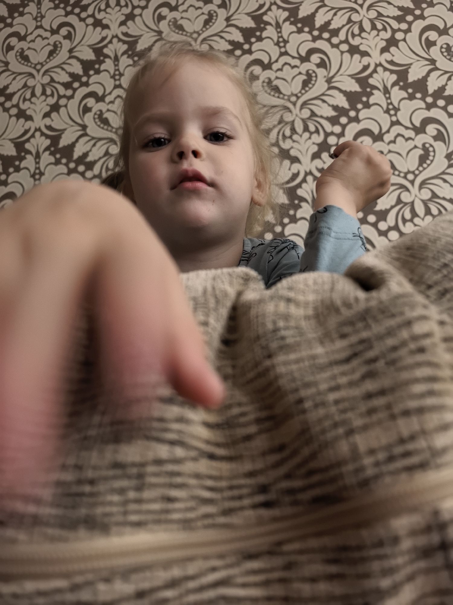 This is a close-up, low-angle shot of a young child with blonde hair. The child is looking directly at the camera with a curious expression. They are wearing a light blue long-sleeved shirt with a dark pattern. Their right hand is raised, and their fist is clenched. To the left of the child, a person's hand, which is out of focus, is reaching towards the camera. The background consists of patterned wallpaper with a cream and brown floral design. A textured, beige and brown blanket is also visible, partially obscuring the child and the foreground hand. The lighting suggests an indoor setting, possibly during the day. The overall scene appears to be a playful interaction between the child and the person taking the photo.