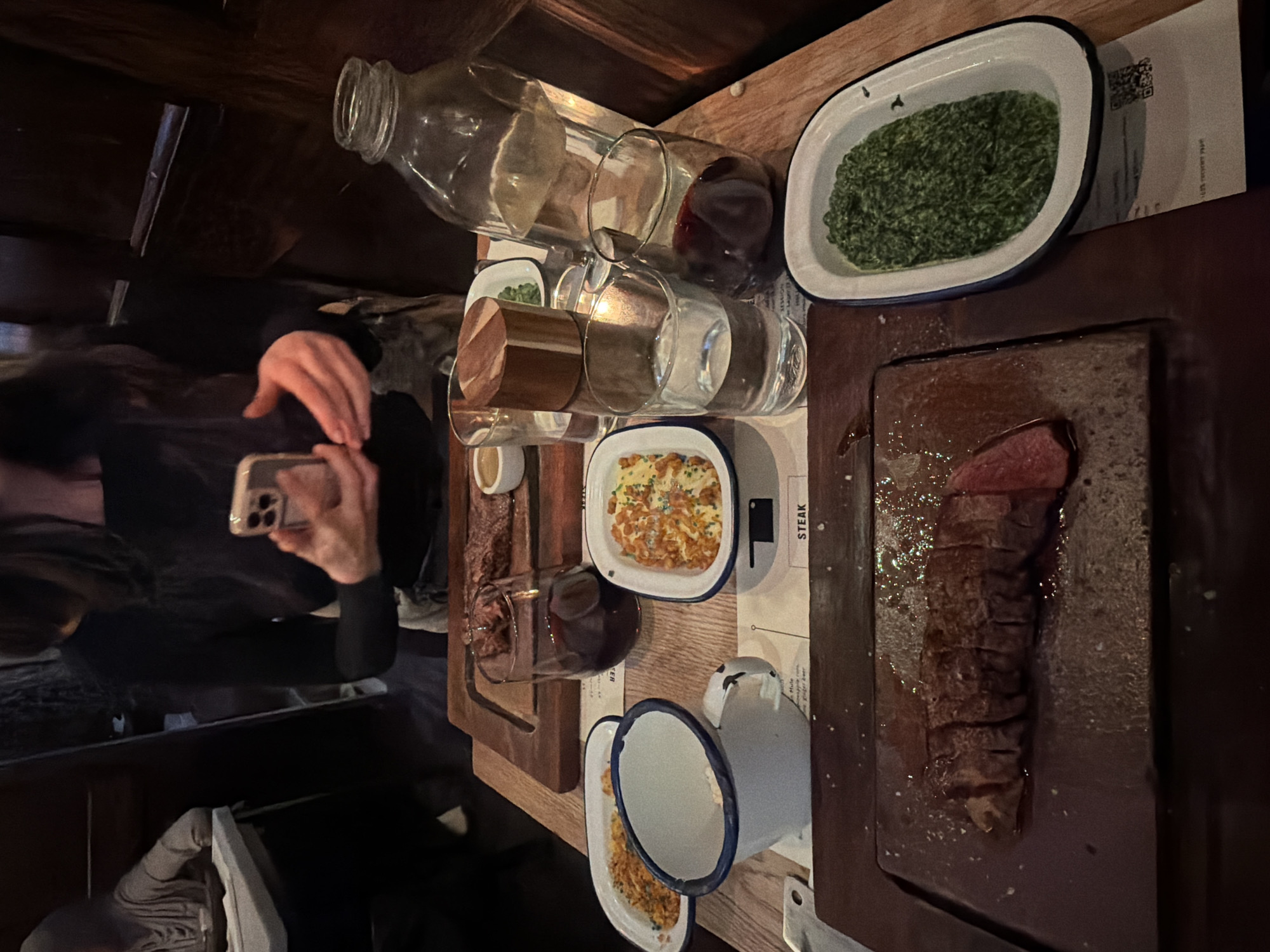 A photograph captures a dining table scene. On the table, there's an assortment of food items including cooked spinach in a white dish with a blue rim, sliced steak on a wooden serving board, and a dish filled with some kind of casserole or baked pasta. Various glassware, including wine glasses, bottles, and what appears to be a ceramic jug, are also present. A person, likely taking a picture of the food with their smartphone, is partially visible on the left side of the frame.
Based on the style of food presentation and the overall ambiance, this photo could potentially be from a restaurant located in the city of London, United Kingdom.