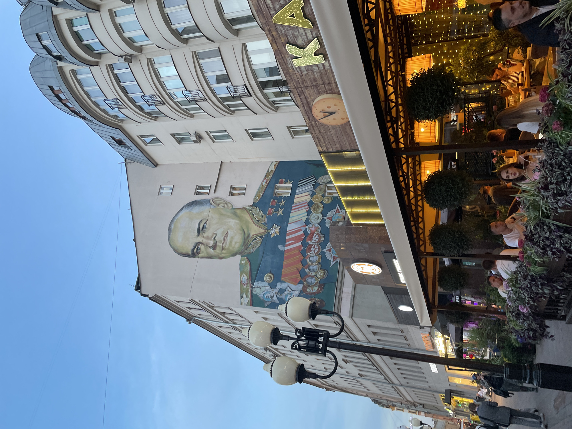 The image shows a street scene in Moscow, Russia. On the left side is a tall building with a mural of Georgy Zhukov, a prominent Soviet military commander, painted on its facade. The building has multiple stories and curved architectural features on its upper floors. There's a clock on the building's facade.
To the right of the building is an outdoor seating area of a restaurant or cafe, with people dining under a decorated awning. The seating area is adorned with plants and lights.
In the foreground, a black lamppost with multiple white globes stands on the sidewalk. People are walking along the street in the background, and storefronts can be seen further down the street. The sky is visible above the building.