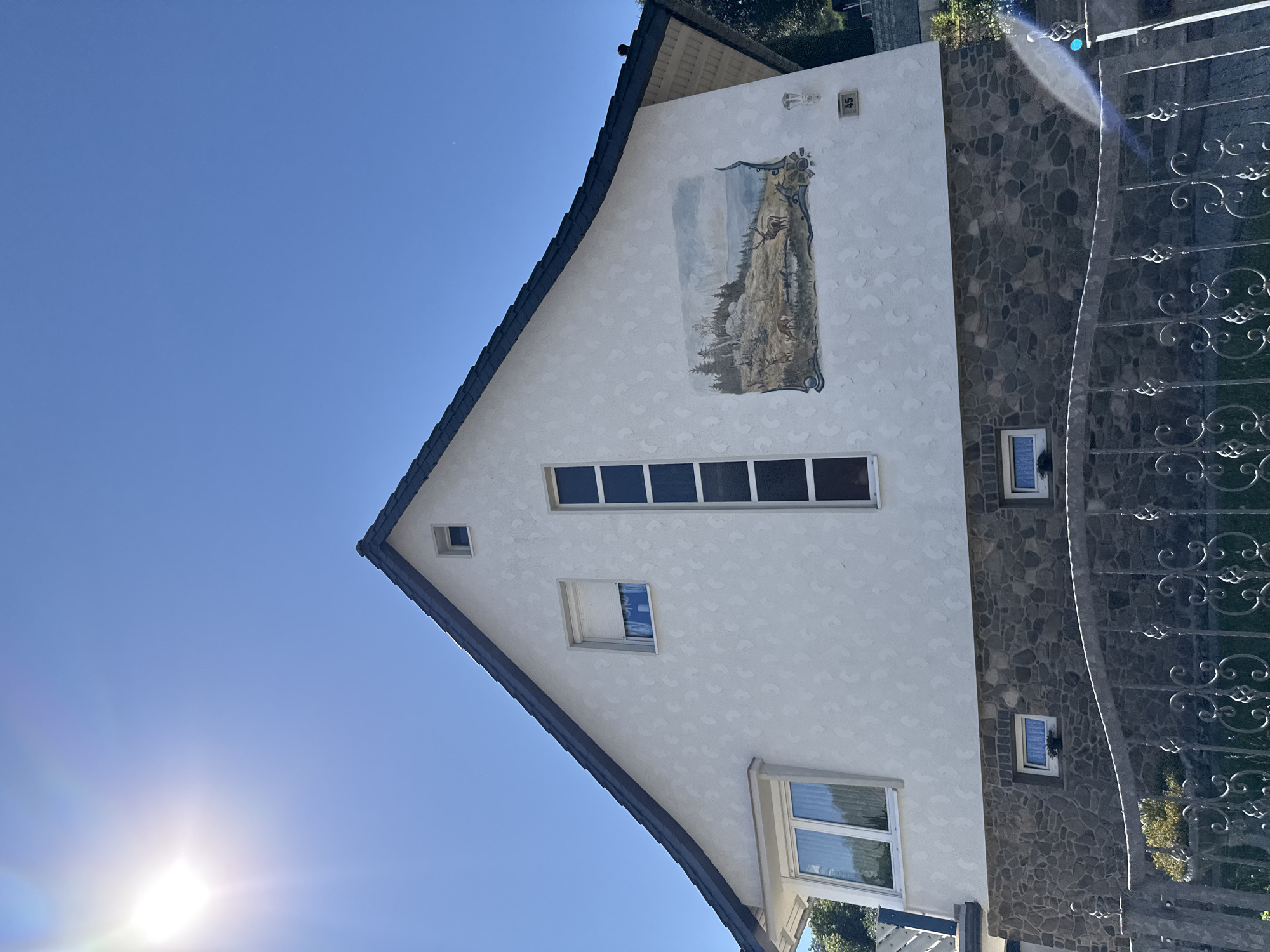 The image shows a two-story house with a dark tiled roof, located on a slope in Bad Lauterberg im Harz, Germany. The upper part of the house is painted white with a textured pattern, and features a mural depicting a forest scene with deer and a stag. There are several windows on this level, including a large bay window on the left and a tall, narrow window composed of multiple panes. The lower part of the house is clad in stone. A decorative metal fence runs along the bottom of the image, in front of a grassy area and some greenery. The sky is clear blue with the sun prominently visible in the upper left corner, casting a lens flare. A house number "45" is visible on the wall near the mural.