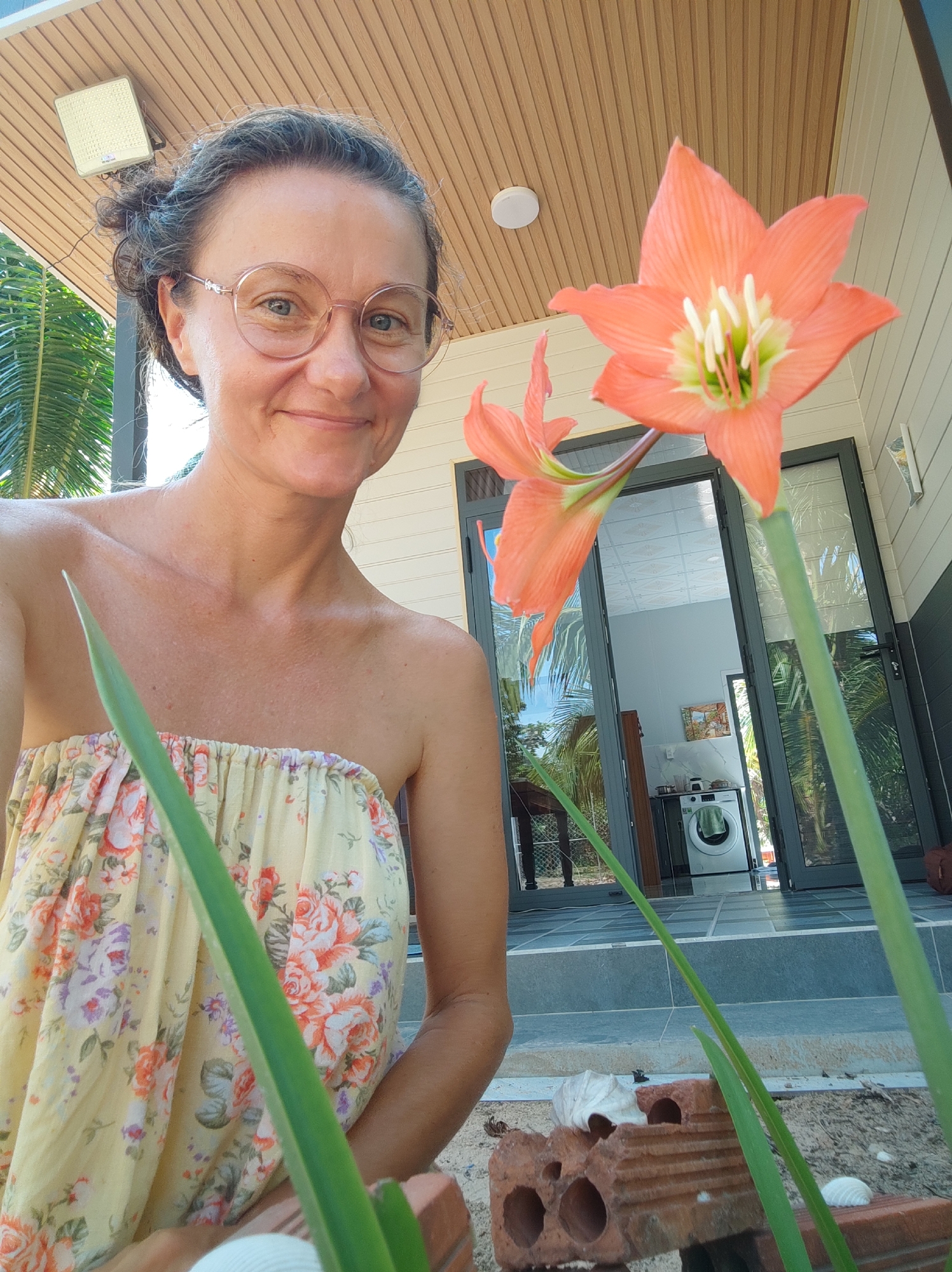 A light-skinned woman with glasses is posing for a selfie outside of a building. She is wearing a floral dress. An orange amaryllis flower is in the foreground. Inside the building, a washing machine is visible. The location appears to be Unknown, Unknown.