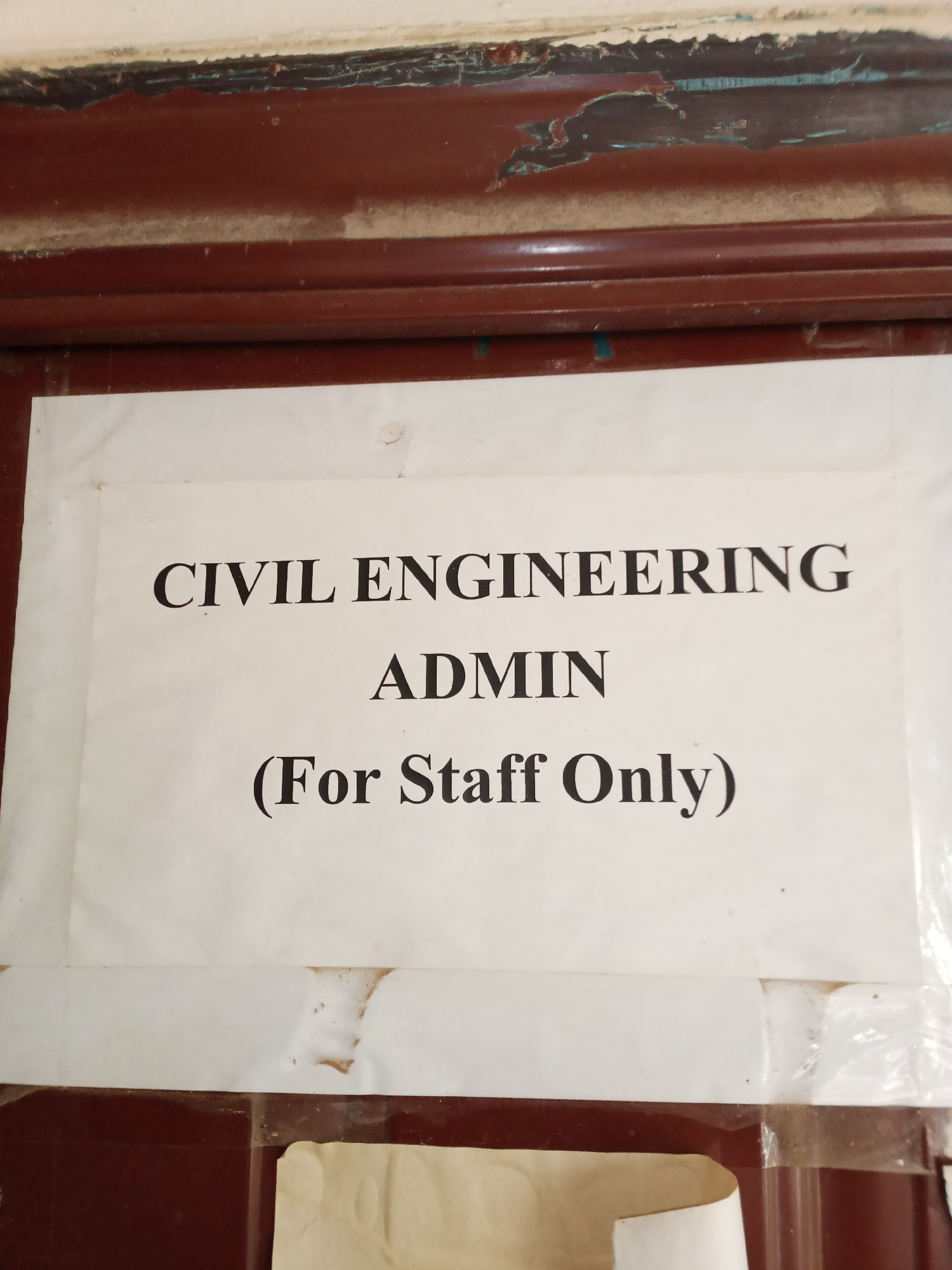A close-up, eye-level shot shows a white piece of paper taped to a dark brown wooden door. The paper has black text printed on it. The top line of text reads "CIVIL ENGINEERING", the second line reads "ADMIN", and the third line reads "(For Staff Only)". The door frame above the paper is peeling and worn, showing layers of brown and a hint of blue underneath. The paper itself has some smudges and discoloration, particularly around the edges. The bottom of the image reveals more of the dark brown door and a corner of another piece of paper, possibly another notice or a flap of paper, also taped to the door. The overall impression is of an office door in a building in Jalingo, Nigeria, indicating restricted access to a specific administrative area within the Civil Engineering department.