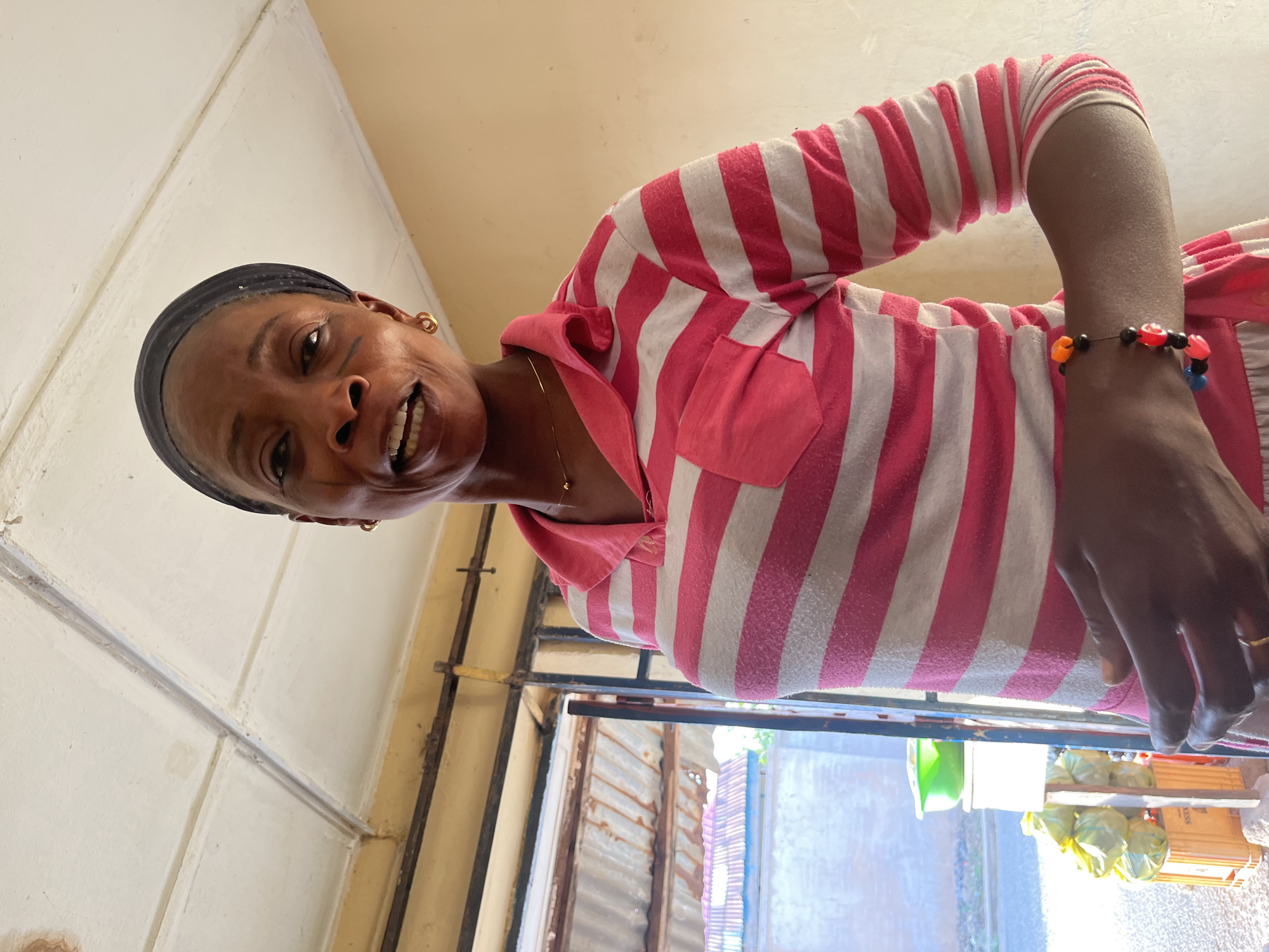 A woman stands indoors, possibly in a residential or commercial space. The location may be Jos, Nigeria.
The woman has dark skin, a black headband, and wears a pink and white striped shirt. She has a beaded bracelet on her wrist and earrings.
The background shows a room with a window, which looks out onto an area with corrugated metal. There is some clutter or merchandise near the window, possibly indicating a shop or small business.