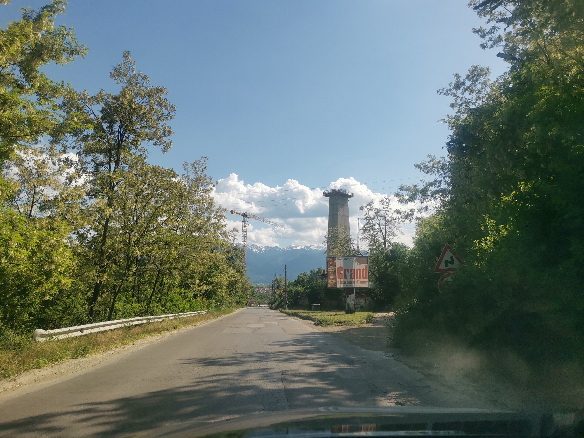 The image is a roadside view in Momin Prohod, Bulgaria. The main feature is a straight asphalt road extending into the distance between verdant trees. A white guardrail lines the left side of the road. In the background, snow-capped mountains are partially visible under a blue sky with scattered clouds. To the right, a large sign advertises "Grand" Restaurant with "pizza bar & dinner" in smaller text. Next to the sign is a tall, unfinished concrete structure topped with scaffolding, and a construction crane stands nearby. A road sign indicating a curve is partially obscured by foliage on the right side. Shadows from the trees fall across the road, and the dashboard of a vehicle is visible in the foreground.
