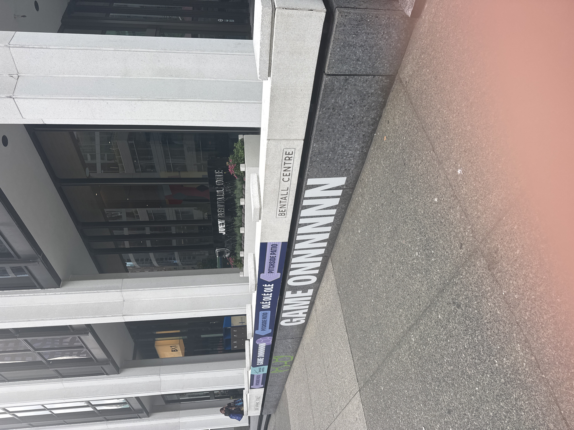 The image shows the exterior of the Bentall Centre in Vancouver, Canada. A long, low concrete planter box runs along the sidewalk in front of the building. The planter box has two levels. The lower, darker level has the words "GAME ONNNNN" written in large white block letters. The upper, lighter level has a series of signs. From left to right, the signs read: "505 BURRARD STREET", "PITCHSIDE PATIO", a purple arrow pointing right, "GAME ONNNNN", a purple arrow pointing right, "PITCHSIDE PATIO", "OLÉ OLÉ OLÉ", a purple arrow pointing right, "PITCHSIDE PATIO". A person is visible in the far left of the image, walking away from the camera. The building behind the planter box has large glass windows and stone pillars. There are some plants and flowers in planters on the upper level of the planter box.