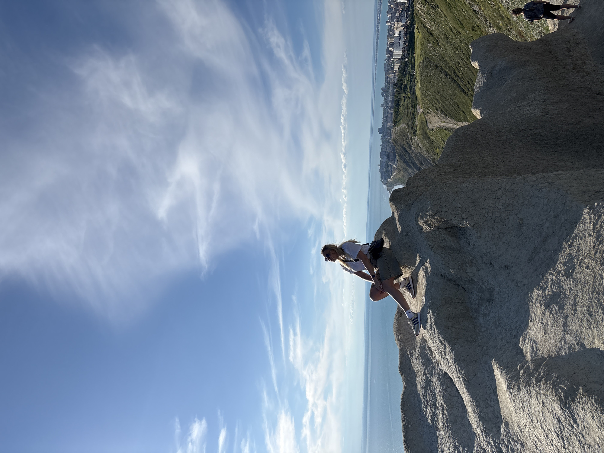 An outdoor photograph captures a woman posing on a rugged, light-colored cliff overlooking a vast coastal landscape. The sky, a clear blue with wispy white clouds, fills the upper left and central parts of the frame, indicating bright, fair weather.
The woman, centrally located on the right, crouches on the rocky terrain, her body angled towards the left. She has long blonde hair, wears sunglasses, a white t-shirt, khaki shorts, and Adidas brand sneakers. A dark bag is strapped across her torso. Beyond the cliff, a calm body of water extends to the horizon, meeting a distant coastline dotted with a modern city skyline and green, undulating hills. Another person stands further up on a grassy slope in the upper right background. The rocky foreground shows intricate textures and shadows, suggesting direct sunlight, likely late morning or afternoon. The scene captures a moment of outdoor activity, possibly hiking or sightseeing, against a picturesque natural and urban backdrop.