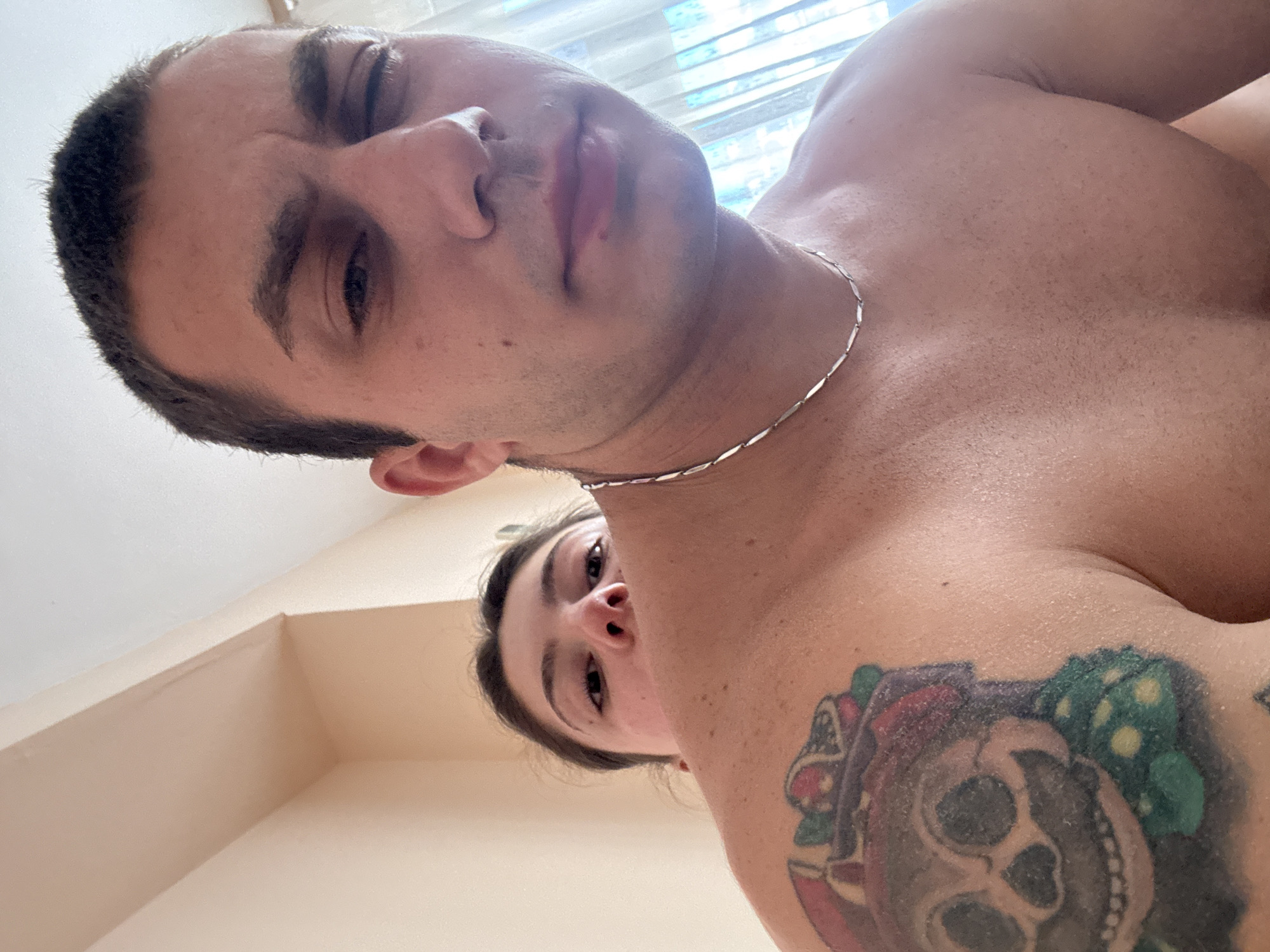 The image depicts a close-up, waist-up view of a shirtless man with a tattoo on his left shoulder. He is wearing a silver chain necklace. Behind him, a woman is visible, her face peeking over his shoulder. The background appears to be an indoor setting with a wall and a curtained window. The lighting suggests it is daytime. The scene does not provide any specific indicators to determine its location within the city of Varna, Bulgaria.