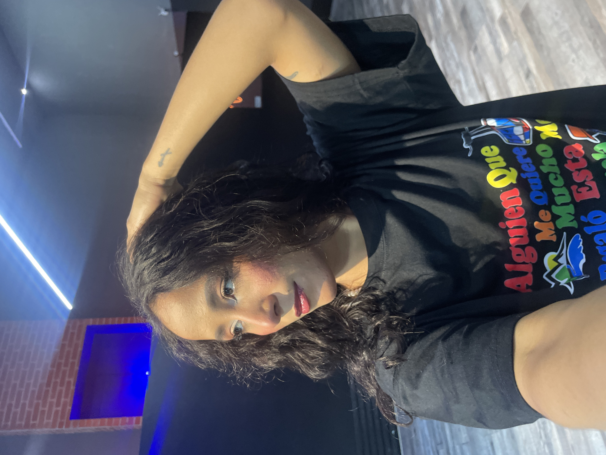 A woman with long, dark, curly hair is captured in a close-up, upper-body shot. She is looking to her left, with her right hand touching her hair. She wears a black t-shirt with colorful lettering and an image of a cable car. The background appears to be an indoor space with dark walls, a brick accent wall with a blue light, and a bright blue light fixture running across the ceiling. The floor is made of light-colored wooden planks. The image quality suggests it was taken with a mobile device. The location of Pampatar, Venezuela is not discernible from the visual elements within the image.
