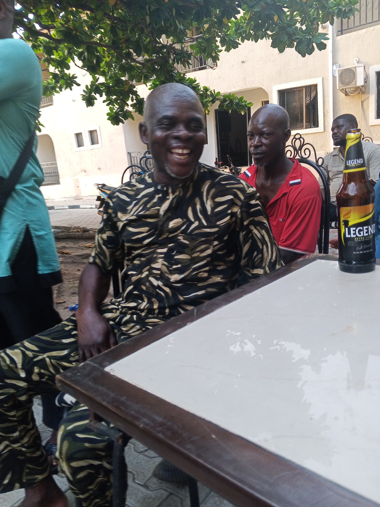 The image shows four men at an outdoor establishment in Lokoja, Nigeria. The man in the foreground is wearing a patterned black and khaki shirt and pants and is laughing. Behind him, a man in a red shirt sits with a serious expression. To the right is a table with a bottle of Legend Extra Stout. The background reveals a building with an air conditioning unit and a man seated behind. Vegetation overhangs the scene.