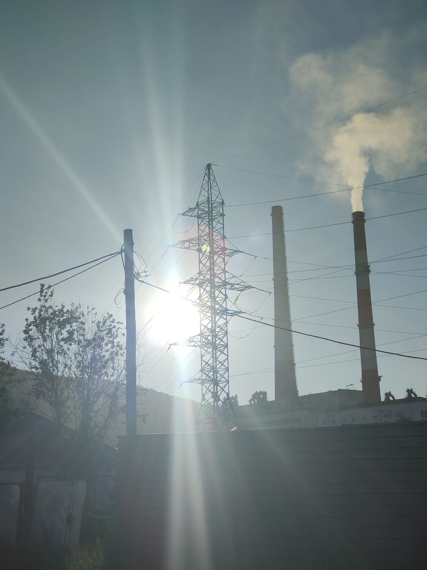 The image depicts an industrial landscape with a power plant in the background, characterized by two tall smokestacks emitting plumes of white smoke. In the foreground, there's a fence, a utility pole with wires, and a transmission tower. The sun is prominently visible, casting strong rays and lens flares across the scene, suggesting it is either early morning or late afternoon. The environment appears to be a mix of industrial and natural elements, with a hint of trees and a hillside visible on the left. The scene is consistent with the industrial infrastructure often found in cities like Magadan, Russia, which is known for its energy production facilities.