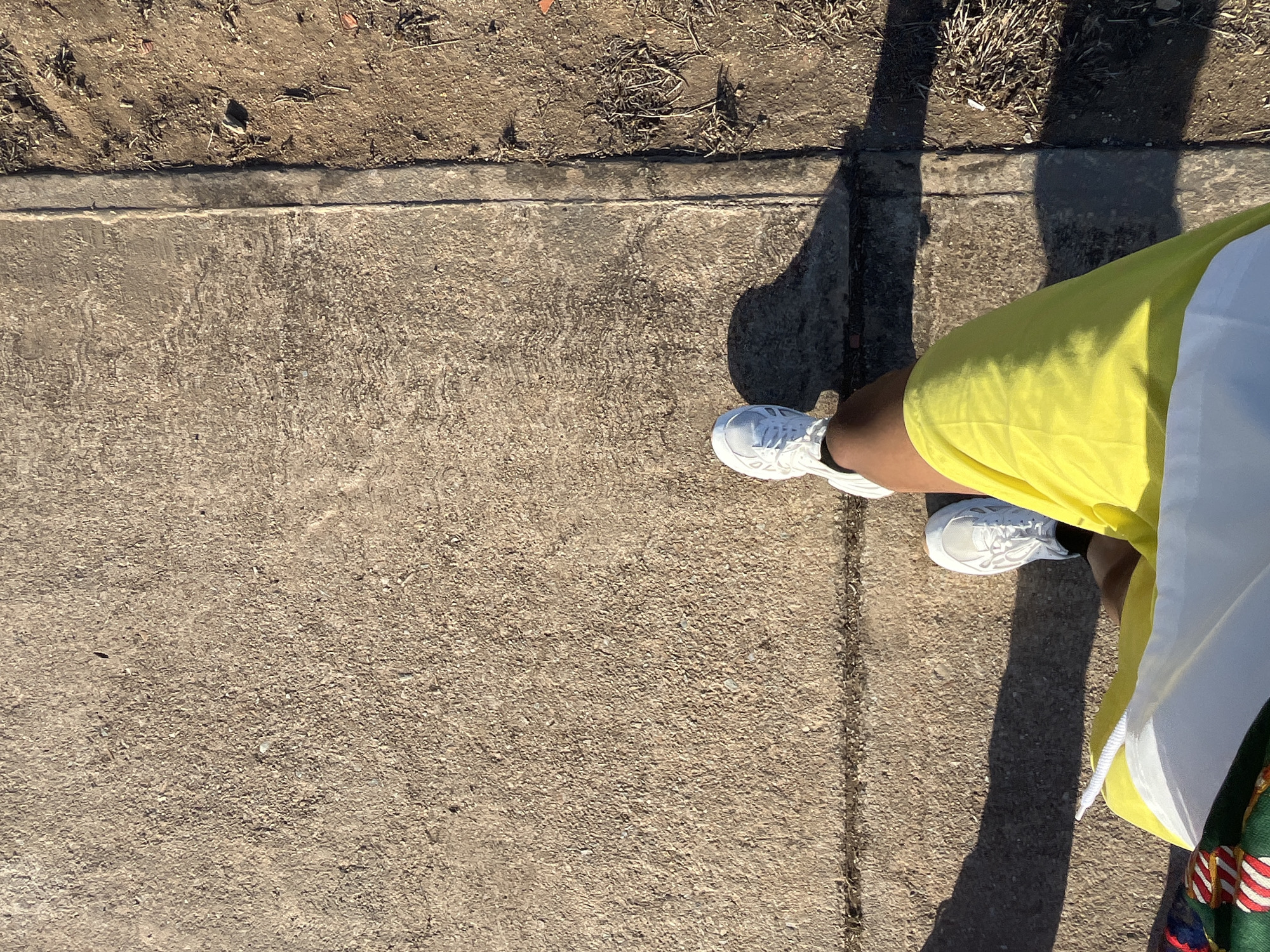 This high-angle, overhead shot captures a person's lower body and the ground. The subject is wearing bright white athletic sneakers with visible laces and soles, paired with vibrant yellow shorts or a skirt. A white garment, possibly a shirt or jacket, is seen partially over the yellow fabric on the right, and a hint of green fabric with red, white, and blue stripes is visible at the very bottom right corner.
The setting is outdoors on a sunny day, indicated by strong, dark shadows cast by the person and other unseen objects, suggesting midday or early afternoon. The ground is primarily a concrete sidewalk with a rough, textured surface. To the upper left, the concrete borders a strip of dry, brown earth containing sparse, withered vegetation and small stones. A visible seam or expansion joint runs horizontally across the concrete where it meets the dirt. The composition focuses on the interplay of footwear, clothing, and the weathered outdoor surfaces under clear skies. No visible text is present in the image. The scene suggests a casual outdoor moment, potentially in Porlamar, Venezuela.
