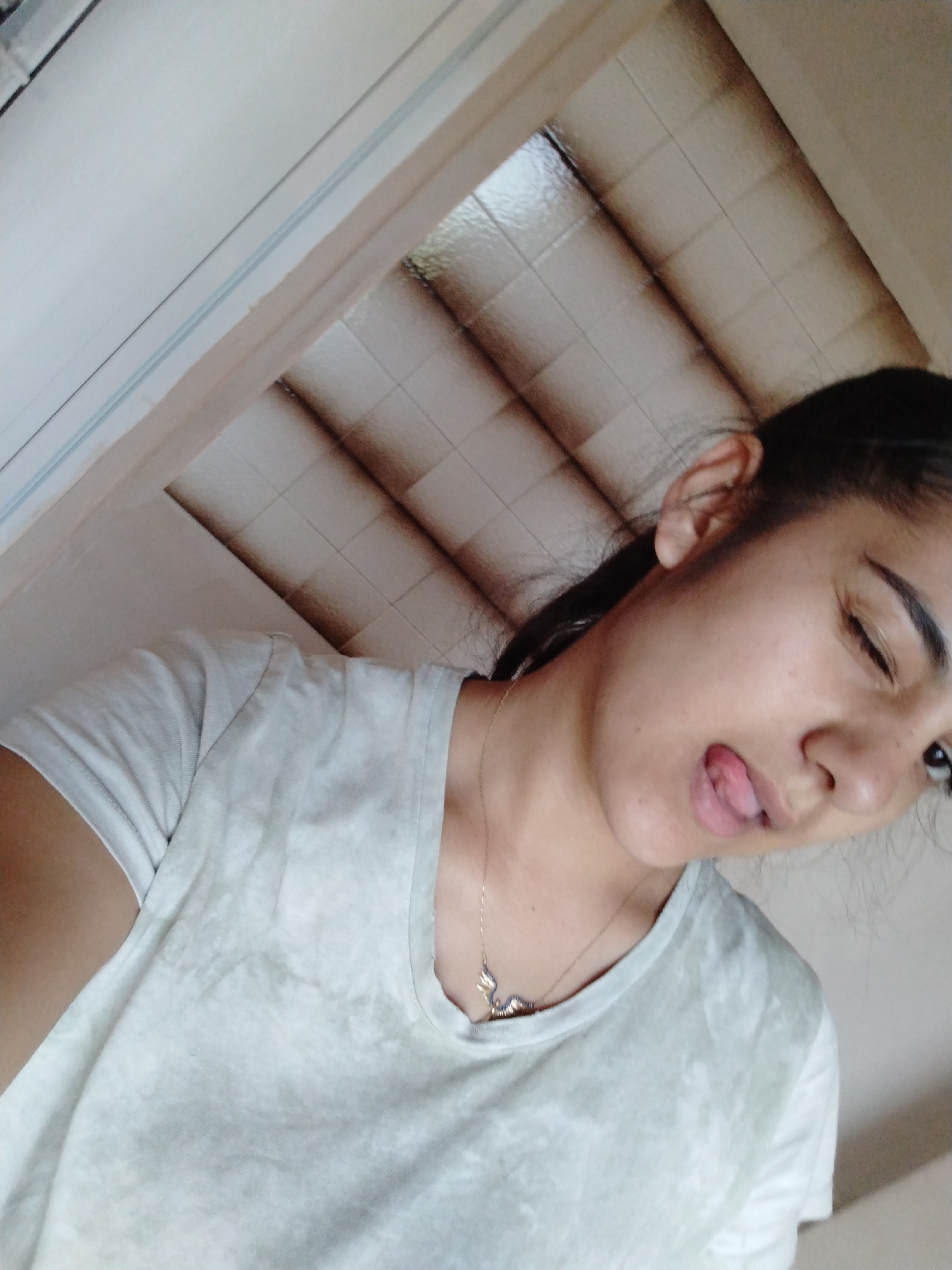 A medium-shot captures a young woman, positioned at an angle, with her head tilted. She has dark hair pulled back into a ponytail. Her left eye is slightly closed, and she sticks out her tongue. A gold necklace with a pendant is visible on her chest. She wears a white t-shirt. The background consists of a white ceiling with tiled panels and a partially visible door frame. The location and city are unknown.