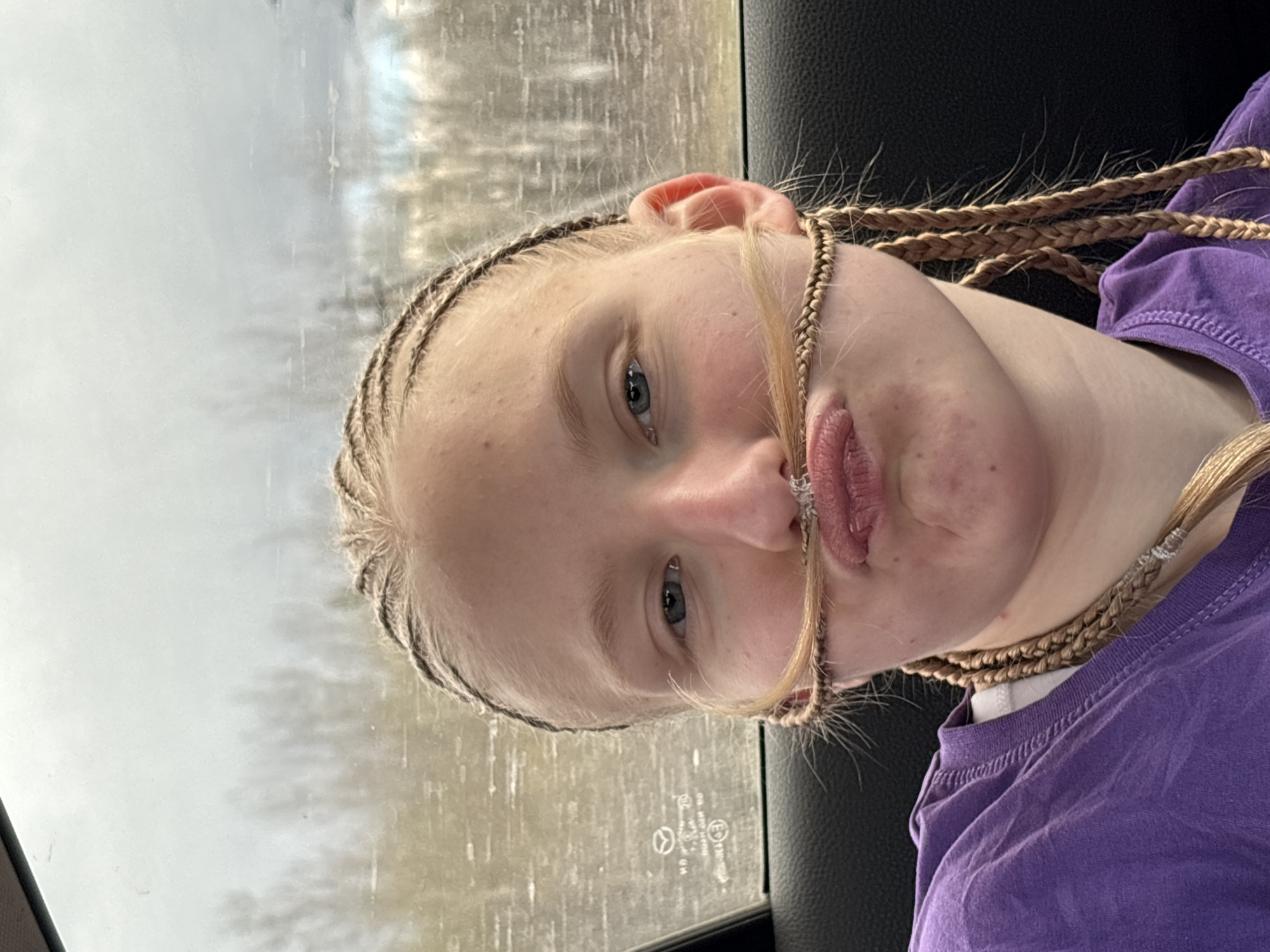 A young, fair-skinned person with blonde hair styled in cornrow braids is depicted in a photograph taken from a slightly elevated angle. The individual is wearing a purple t-shirt and has a strand of hair positioned to resemble a mustache. The subject has blue eyes and is making a pouting facial expression. The photograph appears to be taken from inside a vehicle, with a blurred background visible through the window, possibly depicting a wooded area or overcast sky. The interior of the vehicle, including the seat upholstery, is visible in the frame. The location in the city of Malaya Purga, Russia, cannot be determined from the visible elements of the image.