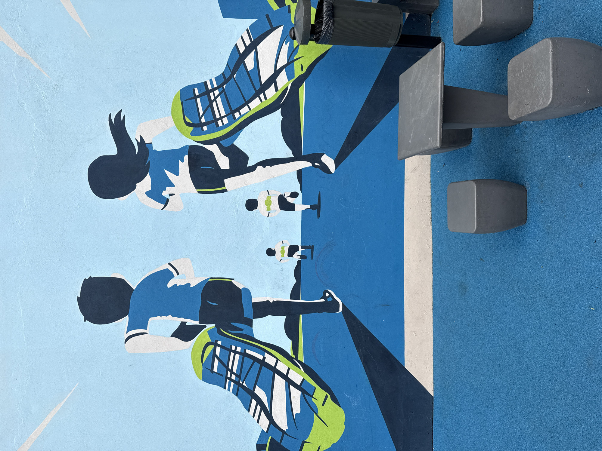 This media file captures an outdoor scene dominated by a stylized mural and real-world urban furniture. The mural, rendered in a flat, graphic style, depicts two large, dynamic figures in athletic attire, along with two smaller cyclists in the background.
The larger figure on the left, possibly male, and the figure on the right, possibly female, are depicted in motion, their bodies angled forward. They wear distinct blue, white, and lime green sportswear, with large, stylized shoes, suggesting participation in a sport like cycling or running. Between these two, two tiny, dark silhouettes of individuals on bicycles are visible, reinforcing the athletic theme. The mural background transitions from a light blue to a darker blue, incorporating abstract shapes and painted shadows.
In the foreground, real objects complement the painted scene. A dark grey trash can with a black liner is positioned near the top right. Below it, on a speckled blue ground, are several grey, block-like structures forming a modern bench or seating arrangement. The ground has a consistent, textured blue finish, resembling a rubberized surface often found in recreational areas. A narrow white stripe runs along the base of the mural.
The scene conveys a sense of energy and athleticism within a public outdoor setting. The location is Buenos Aires, Argentina. No specific time of day or weather conditions are evident. Faint, illegible handwritten text is barely visible on the wall near one of the small cyclist figures.