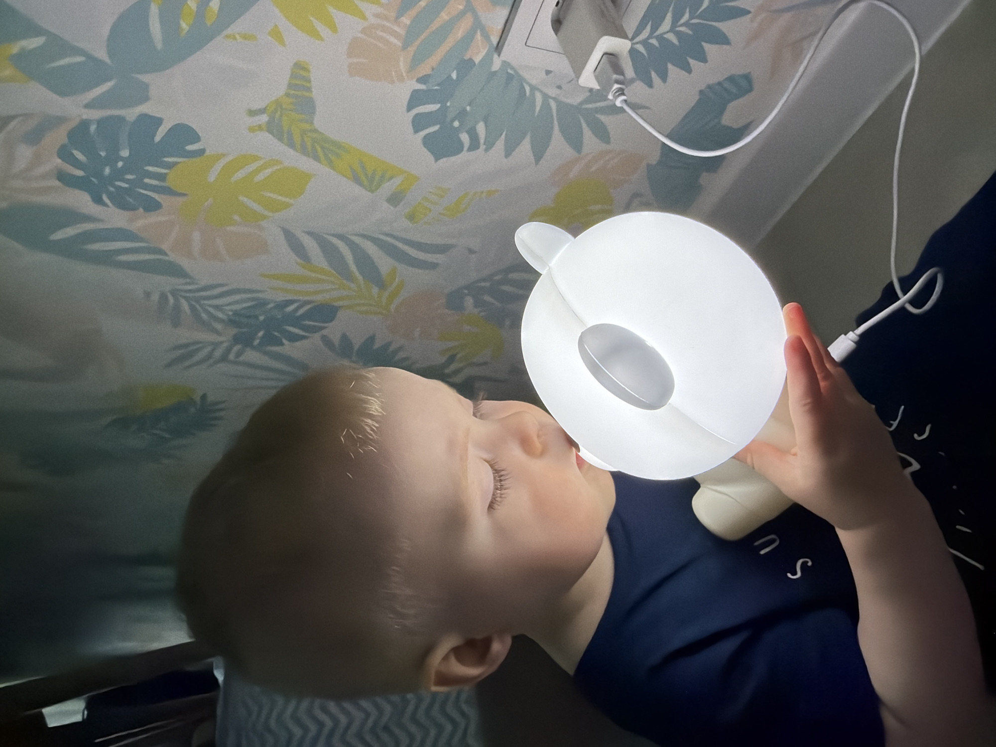 The image shows a close-up, slightly overhead view of a young child in an indoor setting in Votkinsk, Russia. The child, a toddler with light blonde hair, has their eyes gently closed, suggesting they are either sleeping or in a state of calm. They are wearing a dark blue t-shirt with some white, fragmented text or symbols visible, which are difficult to read, appearing as partial letters like 'U' and 'S'.
The child is holding a luminous white night light close to their face. The night light has a rounded shape with two small, visible ears at the top, possibly resembling an animal. A white power cord extends from the light, connecting to a white adapter plugged into an electrical outlet on the wall in the upper right of the frame.
The background features a light-colored wall or textile patterned with stylized tropical leaves, including monstera and palm leaves, in shades of light blue, yellow, and peach. A white shelf or molding runs horizontally behind the child. The primary light source in the image is the glowing night light itself, indicating a dimly lit environment, likely nighttime, which creates a soft and intimate atmosphere around the sleeping or resting child.