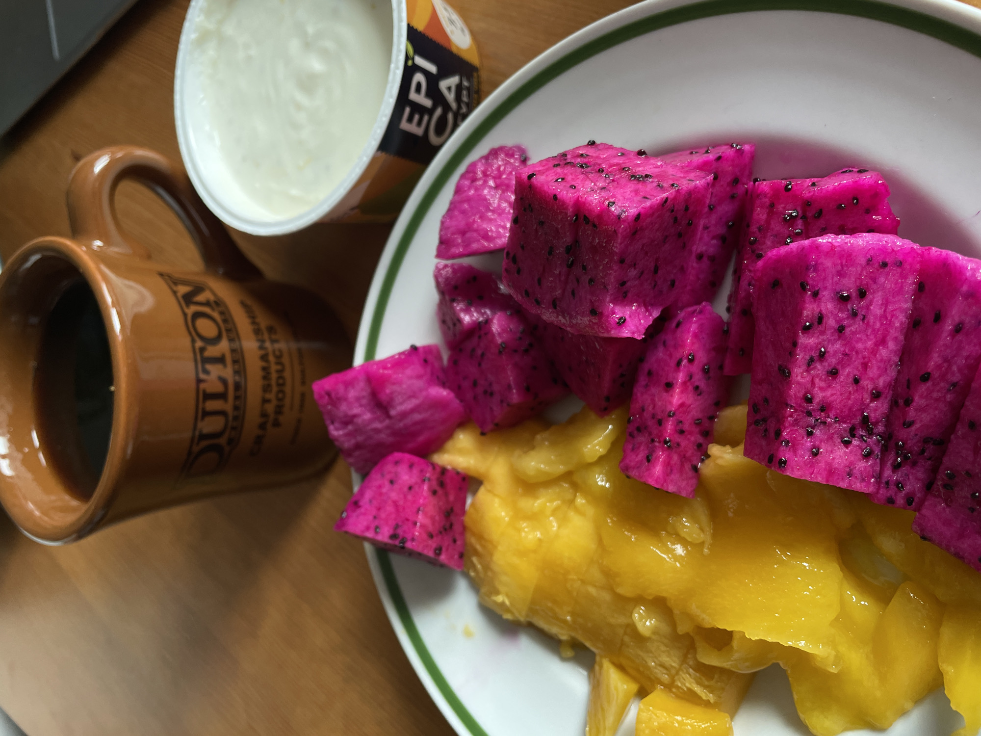 A high-angle, close-up shot shows a breakfast scene. A white plate with a green rim holds cubed pieces of vibrant pink dragon fruit and yellow mango. Next to the plate is a brown ceramic mug filled with a dark liquid, likely coffee or tea. A container of Epica yogurt sits nearby. The items are on a wooden surface. The location and specific city are not identifiable from the image.