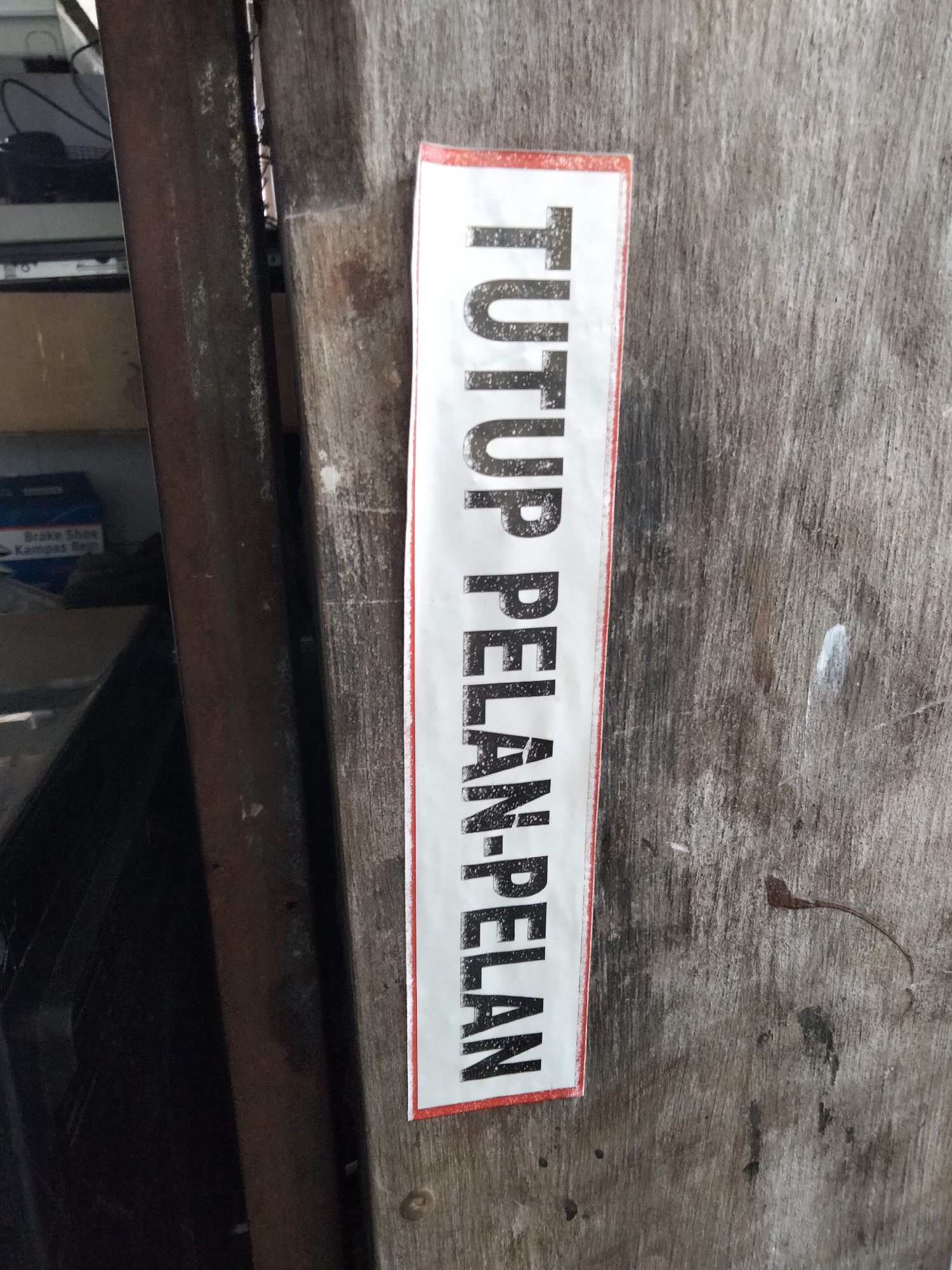 The image shows a white sticker with black text affixed vertically to a weathered, wooden surface. The text reads "TUTUP PELAN-PELAN," which translates to "CLOSE SLOWLY" in Indonesian. The sticker has a thin red border. The wooden surface appears to be a door or a wall, exhibiting signs of age and wear, with visible grain and some dark marks. To the left of the sticker, a dark, vertical metal object is present, partially obscuring a glimpse of shelves and a blue box containing what appears to be automotive parts, specifically "Brake Shoes / Kampas Rem." The overall scene is likely an exterior or interior of a commercial establishment in Denpasar, Indonesia, where such signage is common to indicate careful handling of doors or containers. No people are visible in the image.