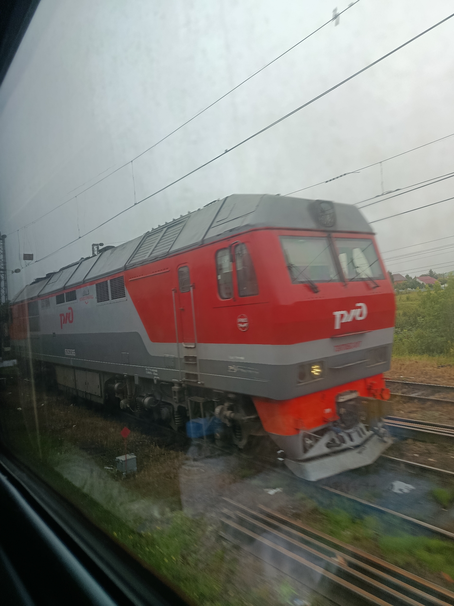 The image shows a red and grey train locomotive moving along train tracks. The train has the logo "РЖД" on its side and front. The locomotive appears to be a modern electric or diesel-electric model. It is a cloudy day, and the train is seen from the perspective of another moving vehicle, likely a train, as evidenced by the reflection of the interior in the window. The surrounding environment includes grassy areas and some trees in the distance, suggesting a rural or suburban setting outside of a city. There are overhead power lines visible above the train, indicating an electrified railway line. The train is in motion, causing some blur in the image, particularly on the wheels and the front of the locomotive. There are no people visible in the image.
