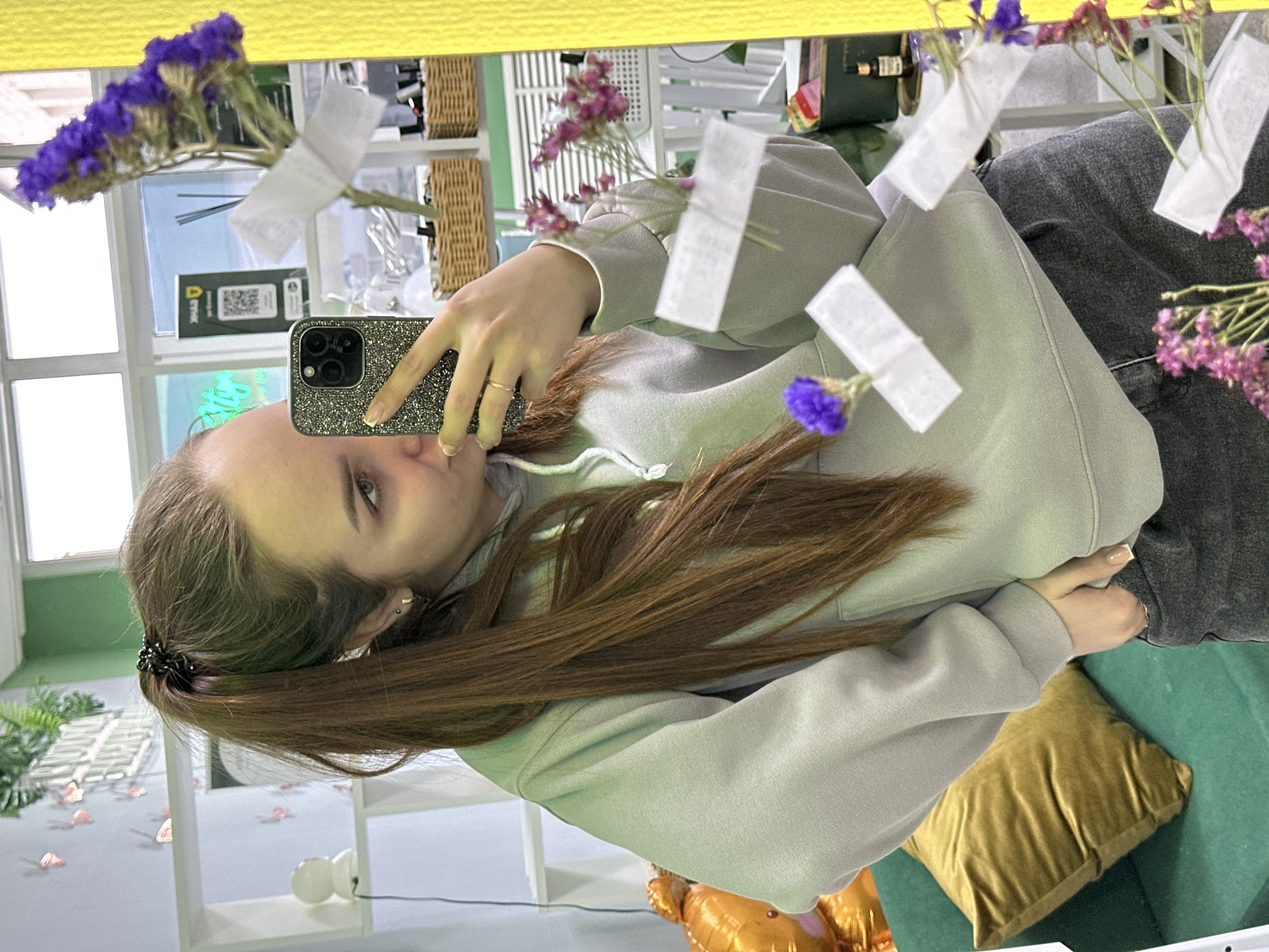 A young woman with long brown hair in a low ponytail is taking a selfie in a mirror. She is holding a glittery iPhone with her left hand. She is wearing a light gray hoodie and gray pants. The mirror is decorated with purple flowers and small white paper notes. The background includes a window, shelving with baskets, a green cushion, and a gold pillow. The location is potentially in Chaikovsky, Russia.