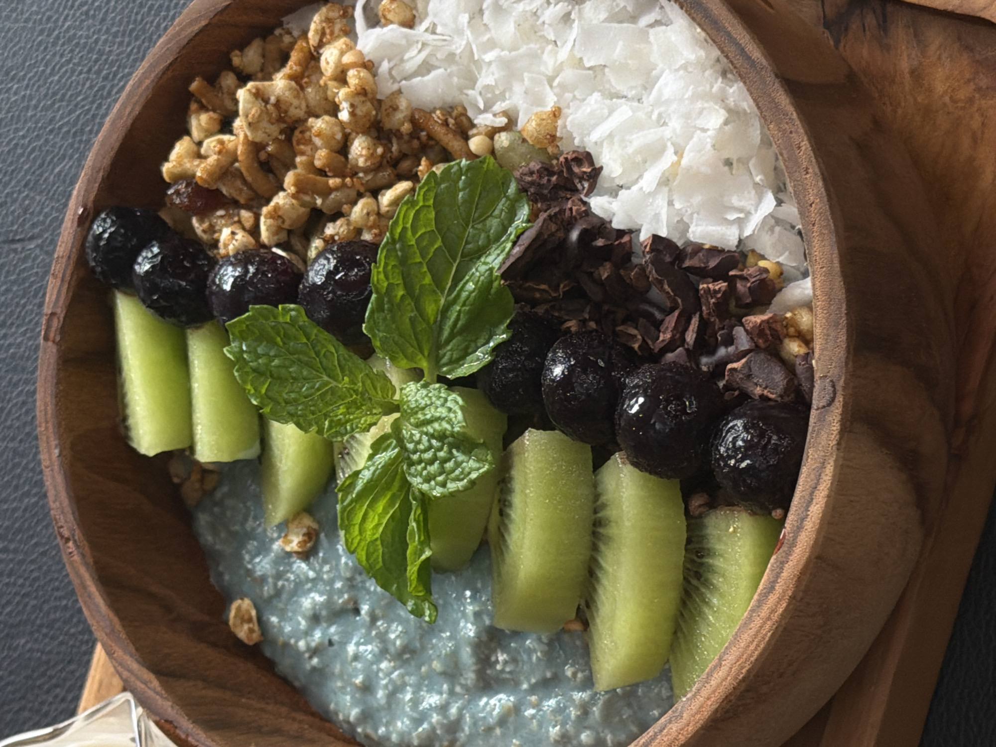 A close-up, overhead shot shows a wooden bowl filled with a colorful and healthy breakfast. The bowl contains a swirl of blue-tinted chia pudding, topped with several bright green slices of kiwi, a cluster of plump blueberries, a sprig of fresh mint leaves, a pile of crunchy granola, and a generous portion of shredded coconut. Dark chocolate shavings are scattered amongst the coconut and granola. The wooden bowl is placed on a dark, textured surface. The setting is a restaurant in North Kuta, Indonesia, as indicated by the ambient lighting and decor typical of establishments in that region.