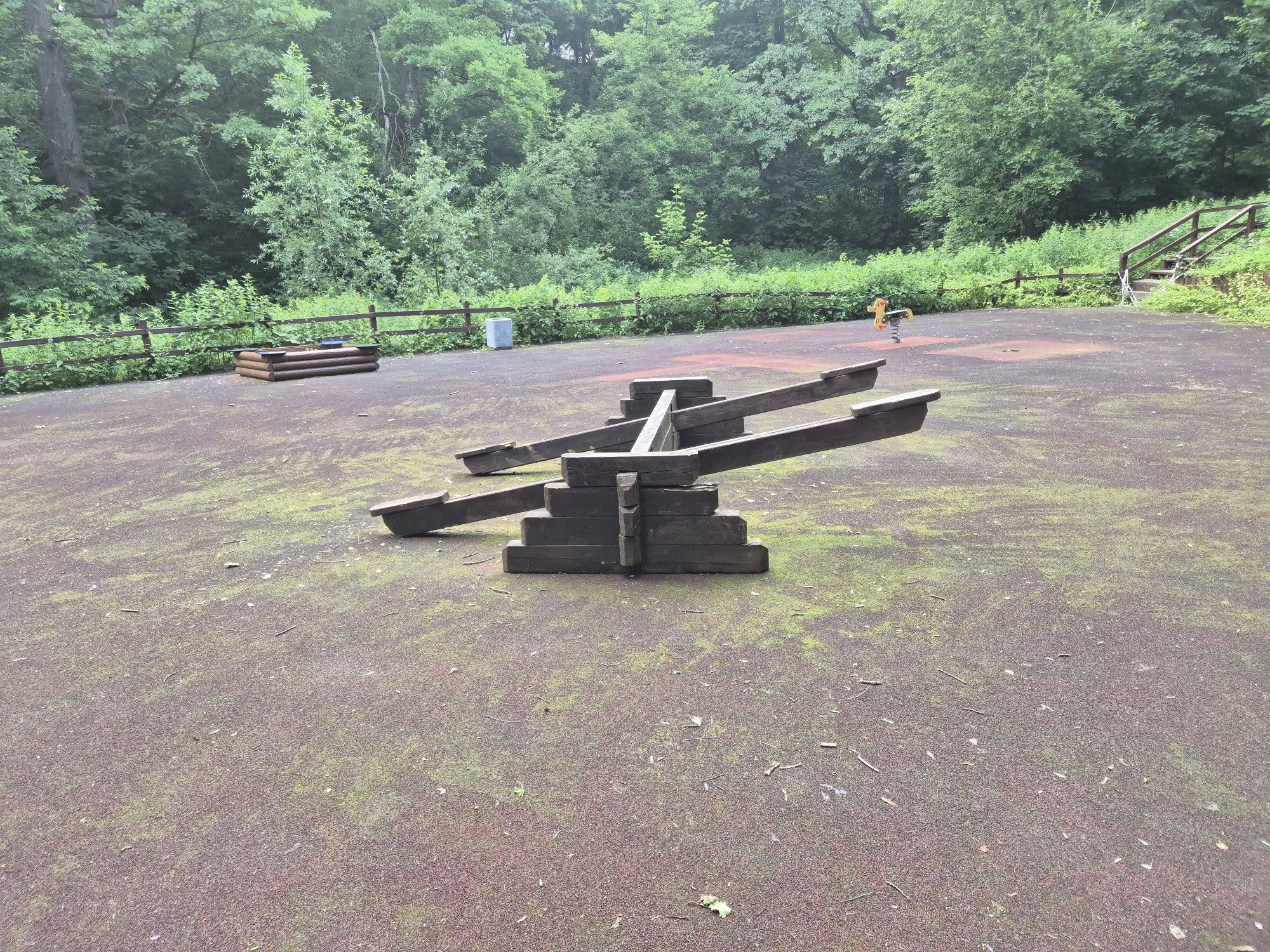 This media file captures an outdoor scene in what appears to be a park or playground, likely in Moscow, Russia. The main subject in the foreground is a wooden seesaw, constructed from thick, dark-stained timber logs stacked to form a sturdy central pivot. The seesaw is positioned on a reddish-brown, textured ground surface that is speckled with green moss and littered with small twigs and leaves, suggesting a natural, somewhat unkempt environment.
In the middle ground, to the left of the seesaw, is a small wooden sandbox. Further to the right, a yellow spring rider in the shape of a horse is visible. Beyond this, a wooden staircase leads up a grassy incline, bordered by a rustic wooden fence. The background is dominated by a dense forest of green trees, indicating a wooded area or parkland. The overall lighting suggests it is daytime, possibly overcast given the diffused light and lack of harsh shadows. The scene evokes a sense of quietness and a slight air of neglect, as the playground equipment appears weathered and the ground is covered in natural debris. There are no people visible in the image, nor is there any discernible text. The atmosphere is peaceful, with nature beginning to reclaim parts of the man-made playground.