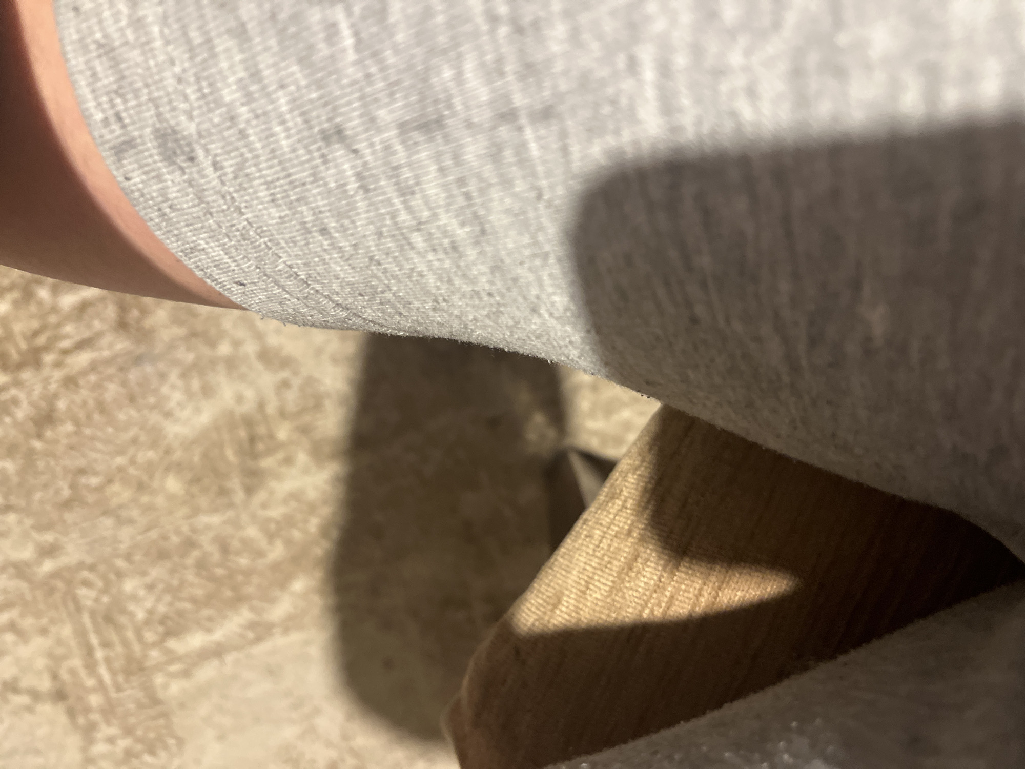 The following is a forensic description of an indoor scene in the city of Blagoevgrad, Bulgaria. The image presents a close-up, high-angle view, showing a segment of a human body, likely a thigh or upper leg, clad in light grey, melange-knit fabric, possibly shorts or sweatpants. The fabric displays a discernible vertical ribbing pattern. Above the fabric, a section of bare, light-tan skin is visible, curving slightly.
To the left of the primary subject, a wooden surface, light brown in color with a subtle horizontal grain, occupies the mid-ground. This object appears consistent with a piece of furniture. The background, particularly in the upper-left quadrant, reveals a light beige or off-white textured surface, possibly a patterned carpet or rug, with indistinct geometric or abstract motifs.
Prominent, soft-edged shadows are cast across the scene. A distinct diagonal shadow bisects the wooden object, and a larger, diffuse shadow extends across a portion of the textured background. Additional shadows delineate the contours between the skin and the grey fabric. Illumination appears to be from an unseen light source positioned to the upper right of the wooden object relative to the camera's perspective, creating well-defined contrast and depth within the frame. No other individuals or specific identifying objects are discernible.